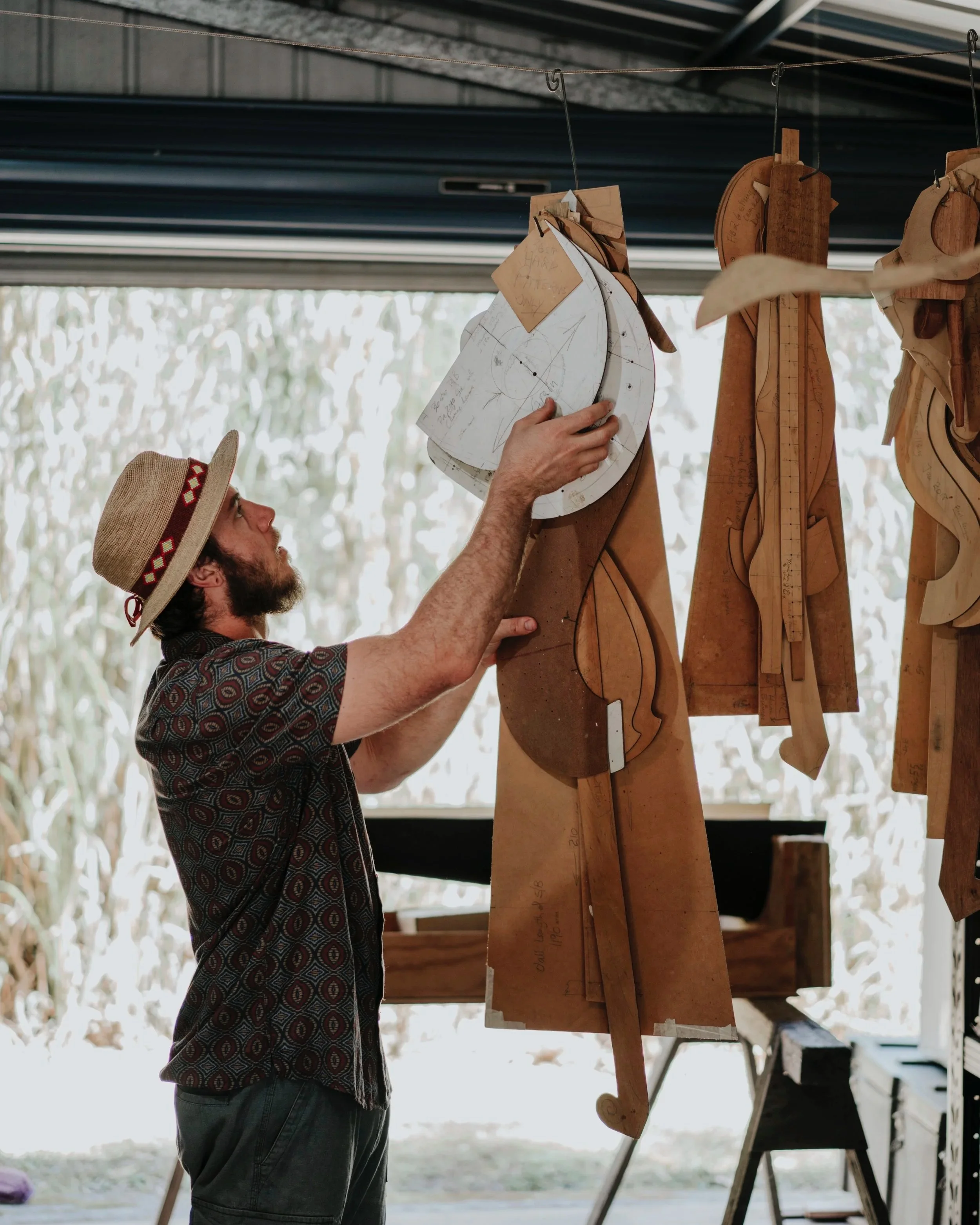 A man wearing a patterned shirt, straw hat, and glasses, working on wooden blacksmithing templates or patterns hanging from a rod in a workshop.