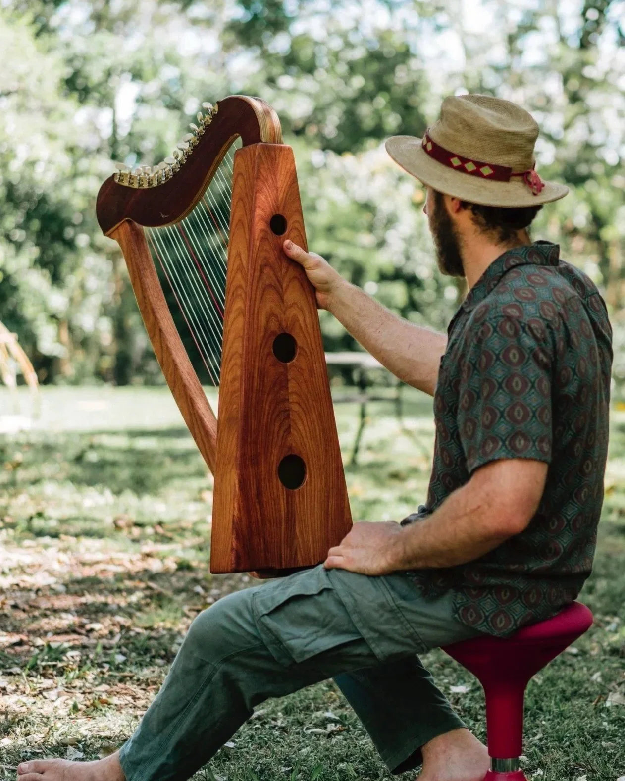 A man wearing a hat, sitting on a red stool outdoors, playing a harp with a wooden frame, surrounded by trees and grass.