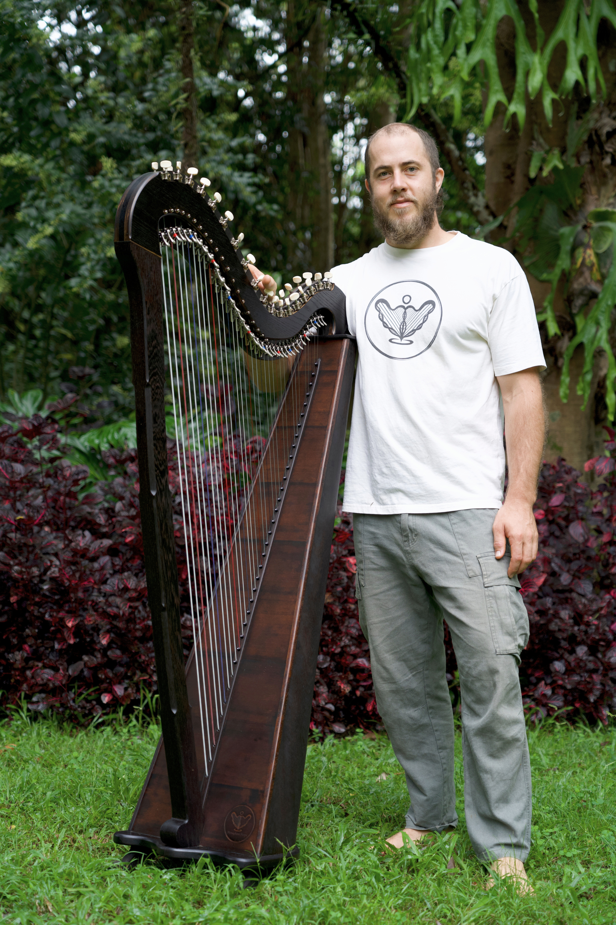 A man standing outdoors on grass with a large harp, surrounded by greenery and bushes.