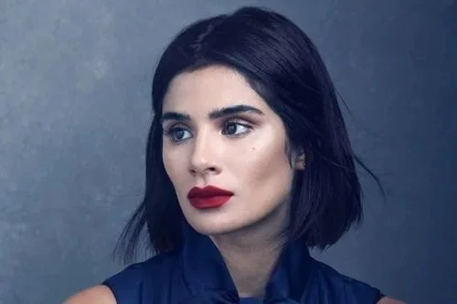 A young woman speaking at a podium during a conference or event, with a microphone and large screen in the background. F4GF Diane Guerrero