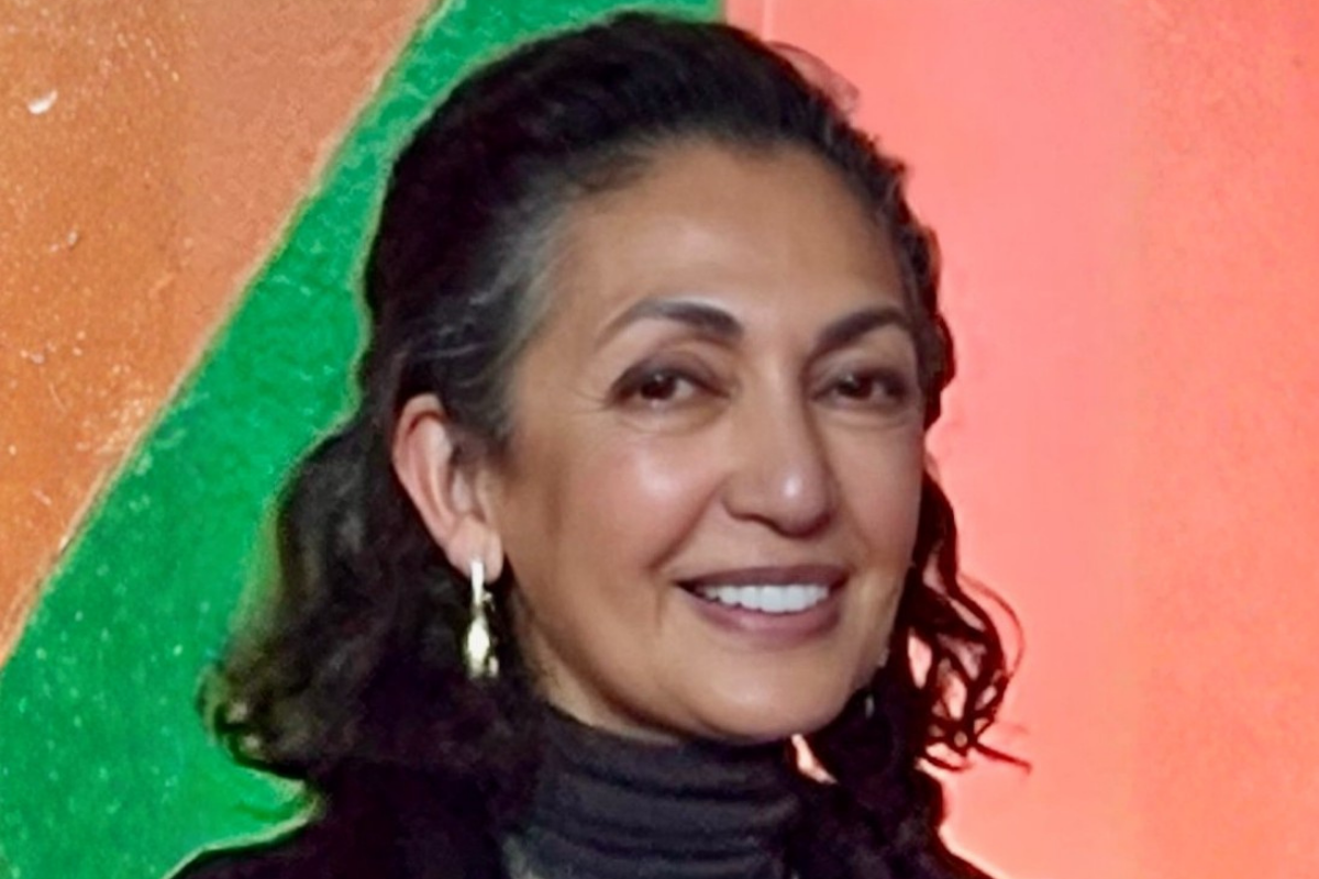 A woman with long dark hair smiling, wearing a turtle neck in front of a colorful background