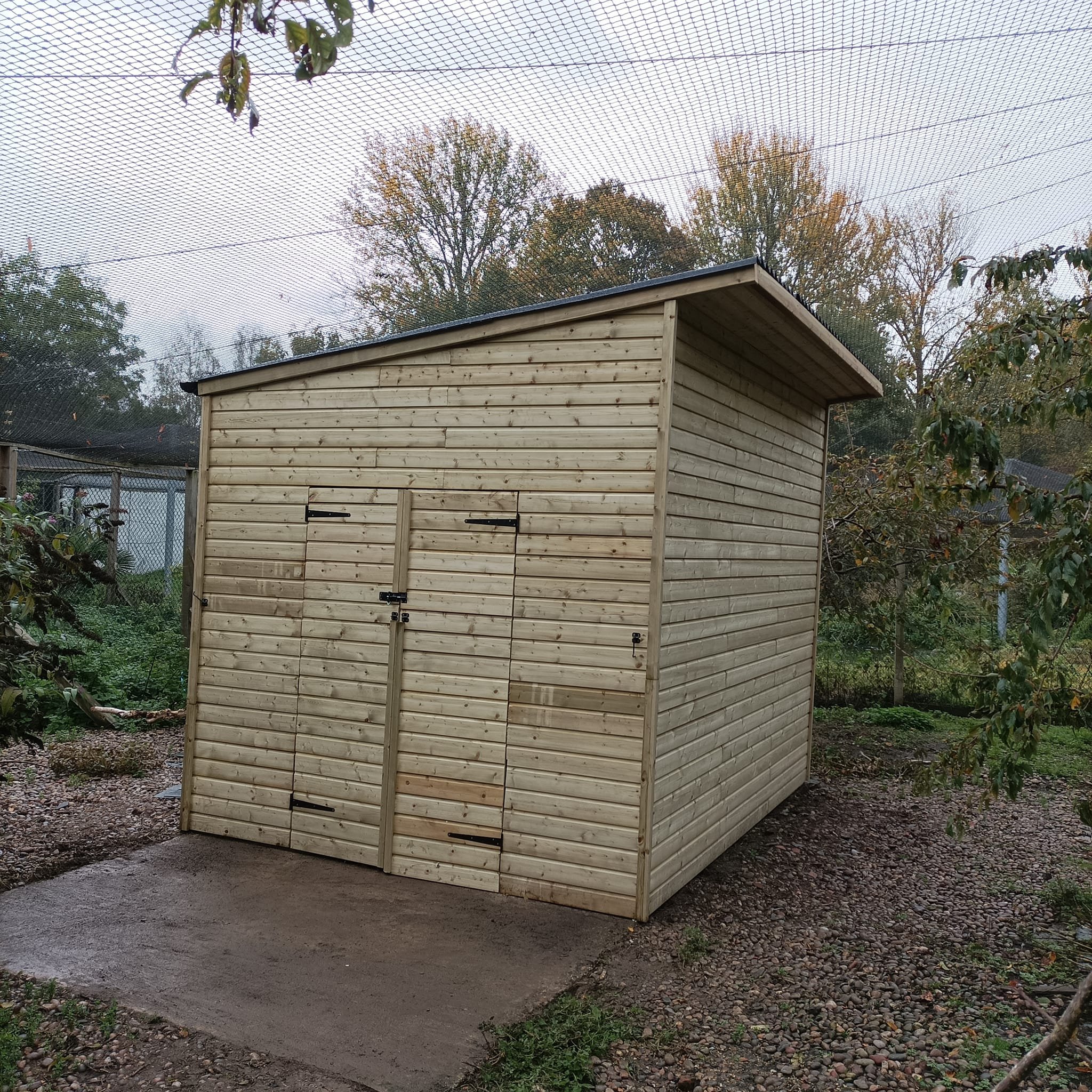 Wooden garden shed with double doors and a slanted roof, situated outdoors in a garden area.