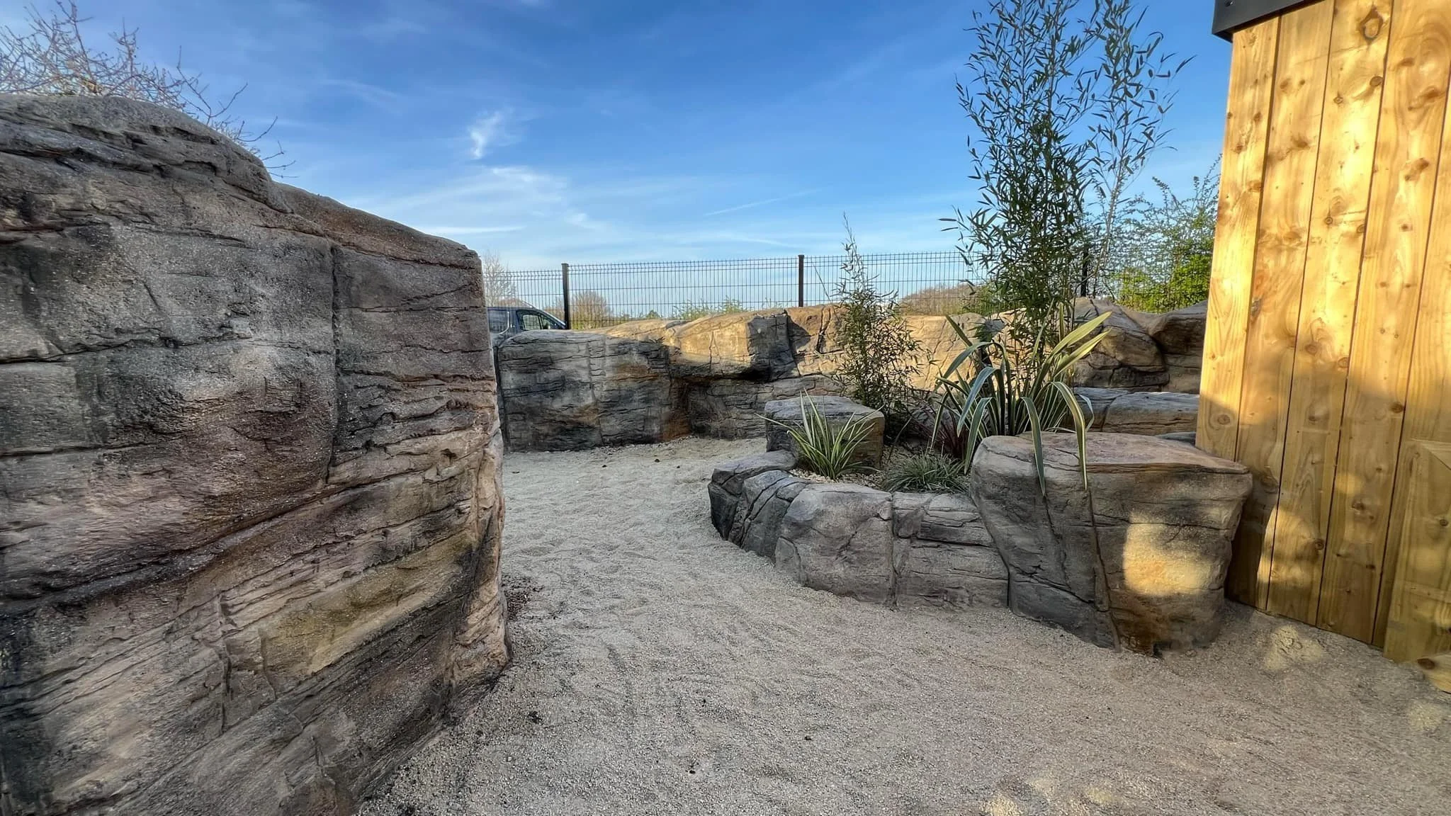 A sandy outdoor space with large rocks and a small plant bed containing shrubs and tall plants, bordered by a wooden fence and a stone wall, under a blue sky with some clouds.
