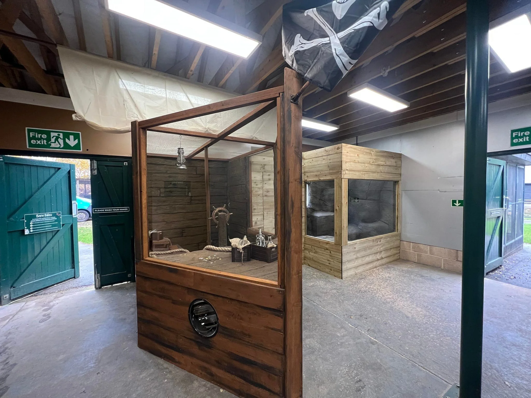 Indoor space with two green fire exit doors, one open, and wooden display structures, including a glass-enclosed display with a wooden frame and another smaller wooden enclosure, with fluorescent lighting and a concrete floor.