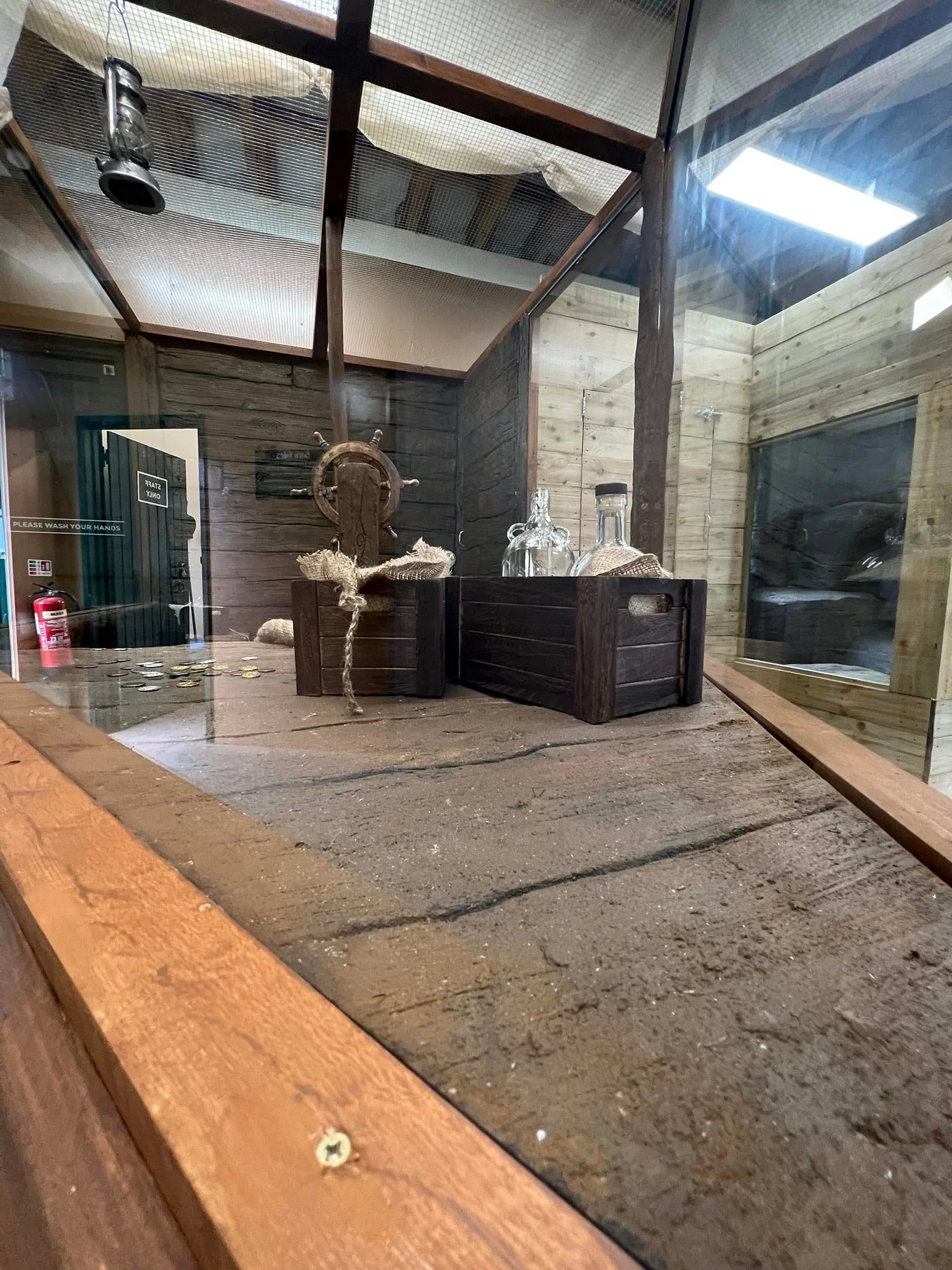 A display case with a wooden top contains a ship's wheel, glass bottles, and coins, with a wooden-paneled room and ceiling in the background.