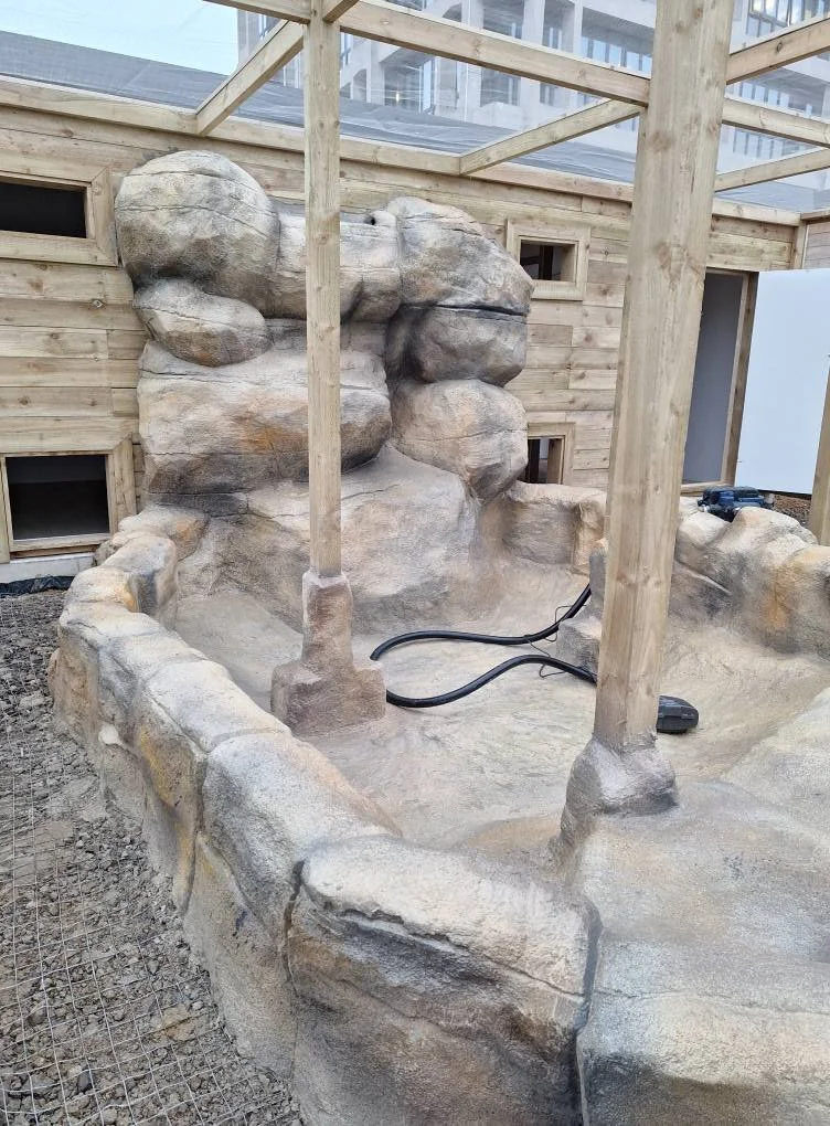 A construction site with a rock formation and a partially built wooden structure. The rock formation resembles a small waterfall or cliff face, with wooden beams and small windows in the background.