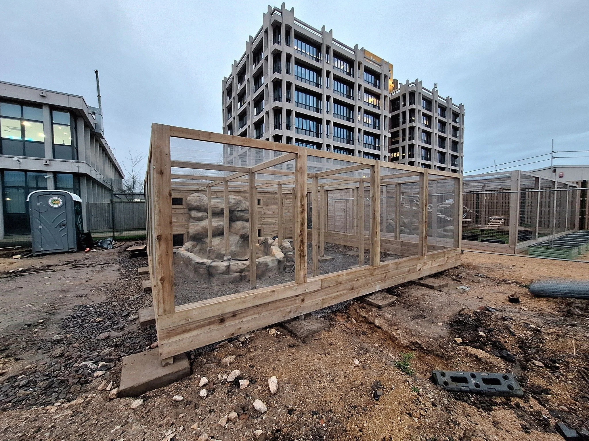 Construction site with a wooden-framed enclosure and an artificial rock formation, with a large multi-story building in the background.