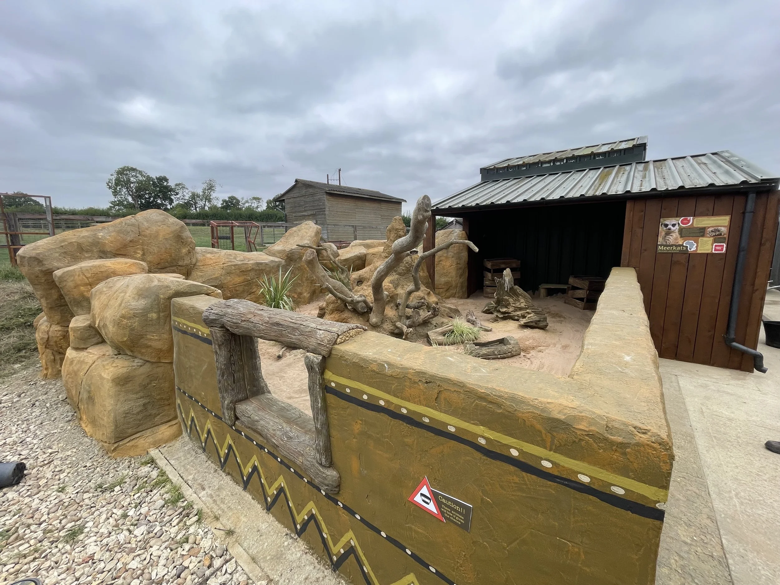A petting zoo area with large rocks, wooden logs, and a small shelter with a sign about meerkats. The enclosed area has a sandy ground and is decorated with Africa-themed art on the shelter wall.