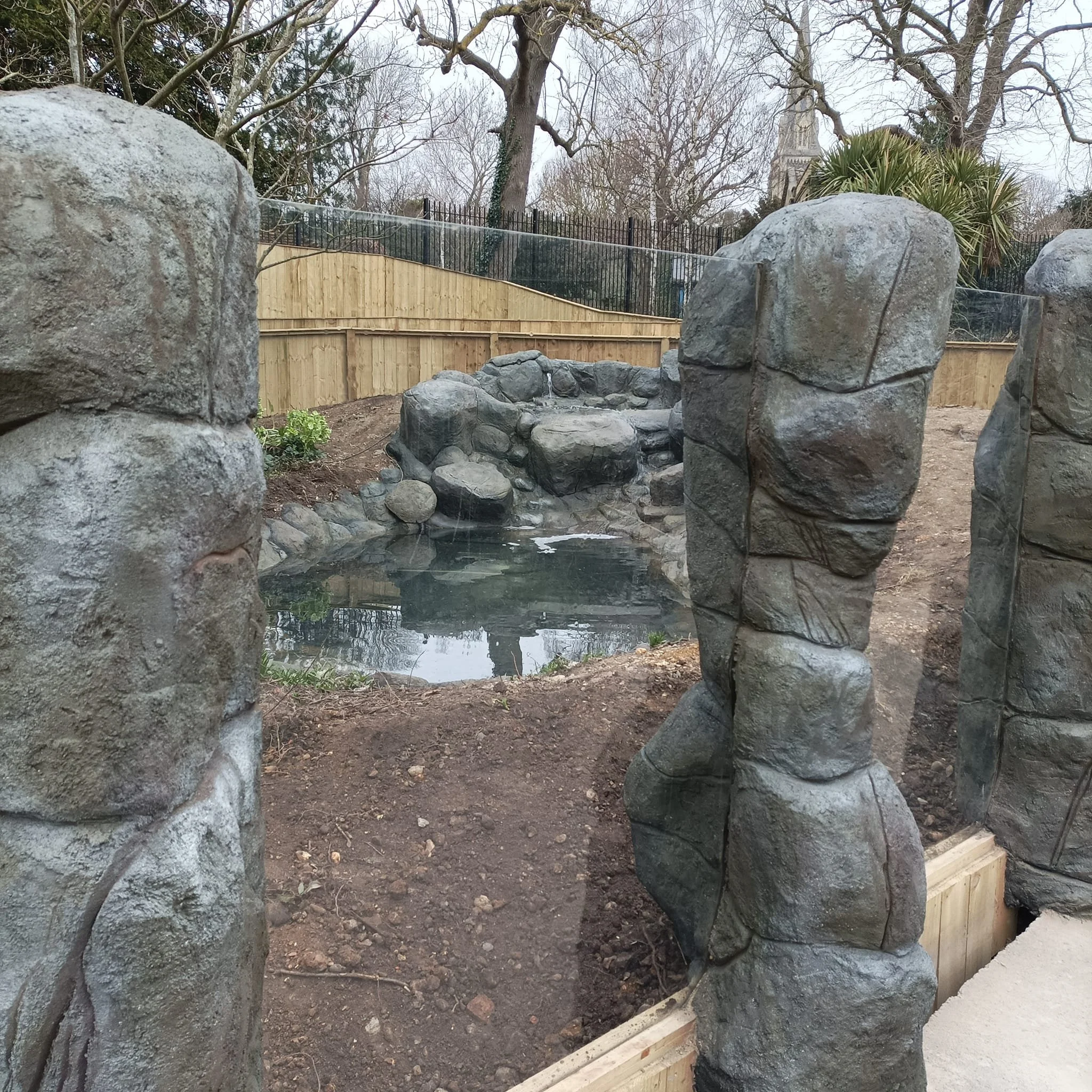 A small artificial pond surrounded by rocks and rocks walls, with trees and a wooden fence in the background.