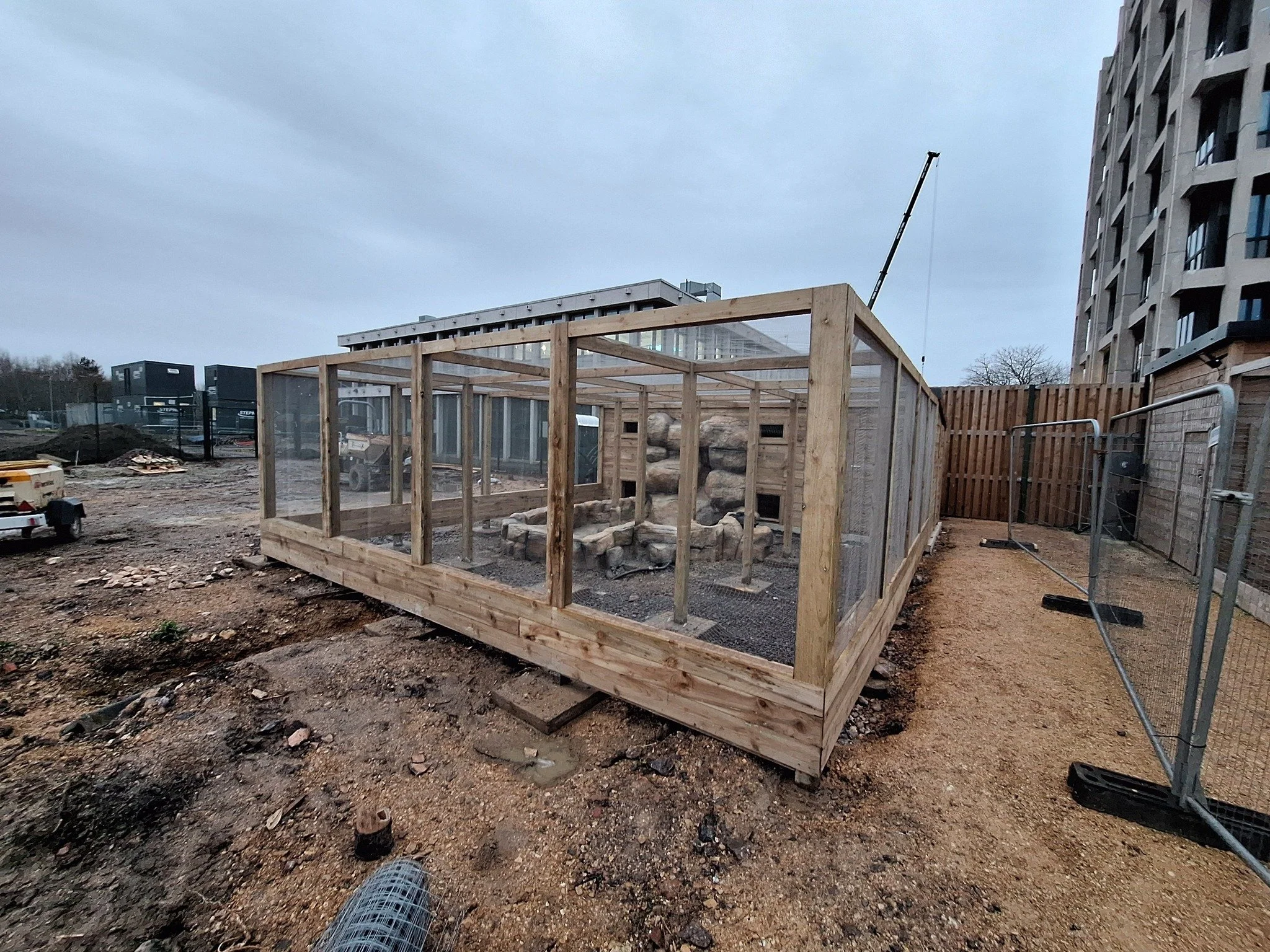 Wooden and mesh enclosure under construction on a construction site, with rocks inside, surrounded by dirt and construction materials, and a high-rise building in the background.