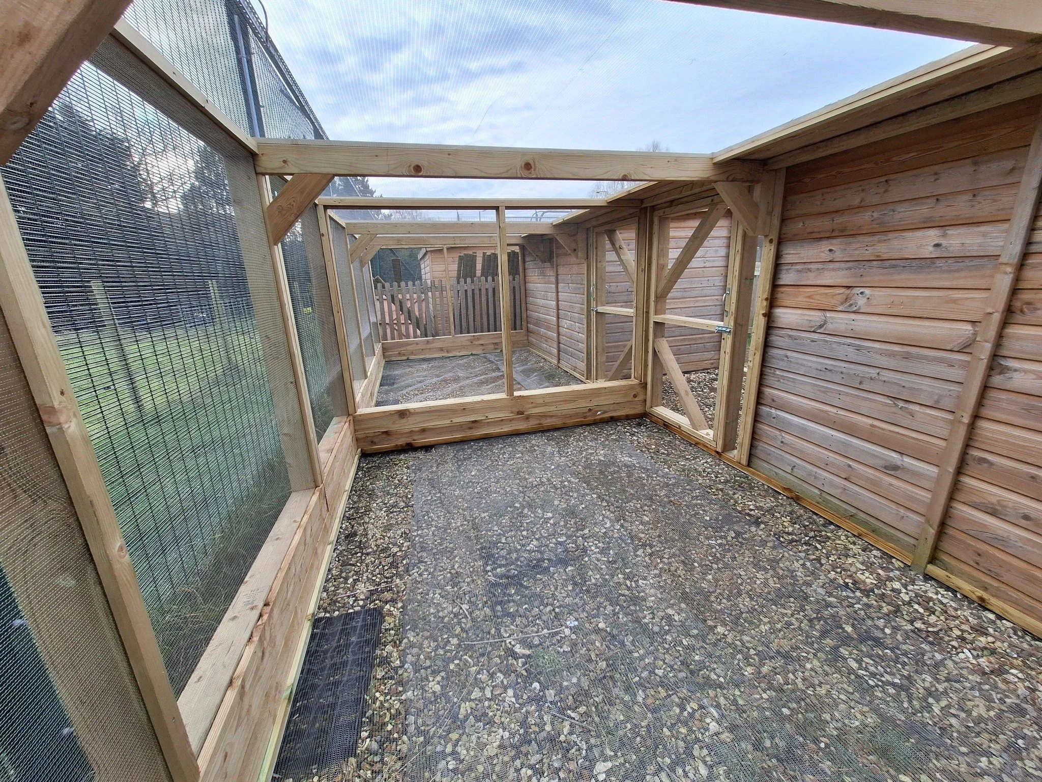 Photo of a wooden chicken coop with a gravel floor, adjacent to a wooden fence and a grassy yard, under a partly cloudy sky.