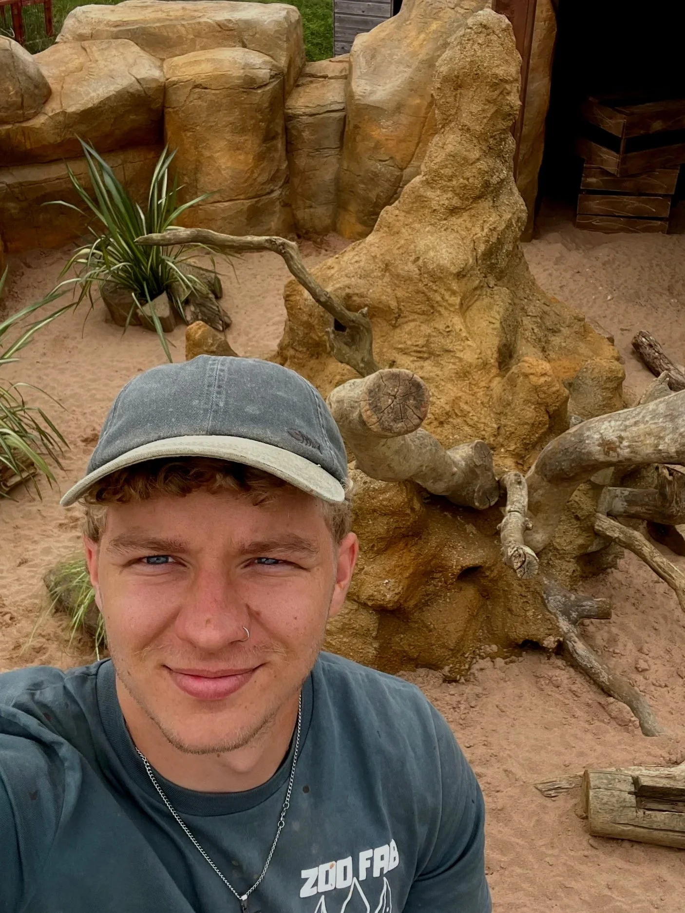 A young man wearing a gray cap and a dark gray Zoo Fauna T-shirt taking a selfie with a large horizontal fallen tree and desert-style rocks in the background.