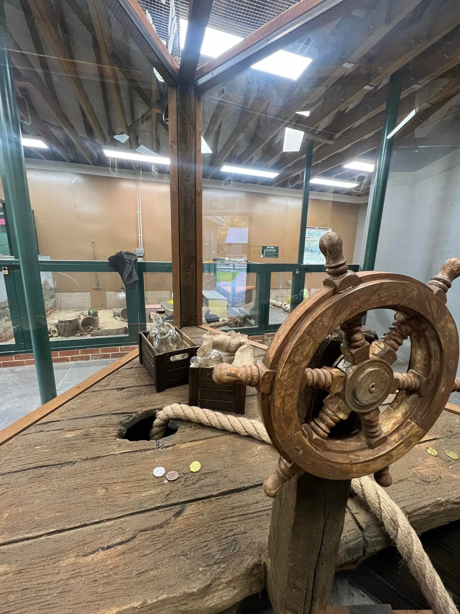 Close-up of a wooden pirate ship wheel on a weathered wooden surface, with a thick rope, glass bottles, and small coins. Behind the glass, there are enclosures with animals in a zoo or park setting.