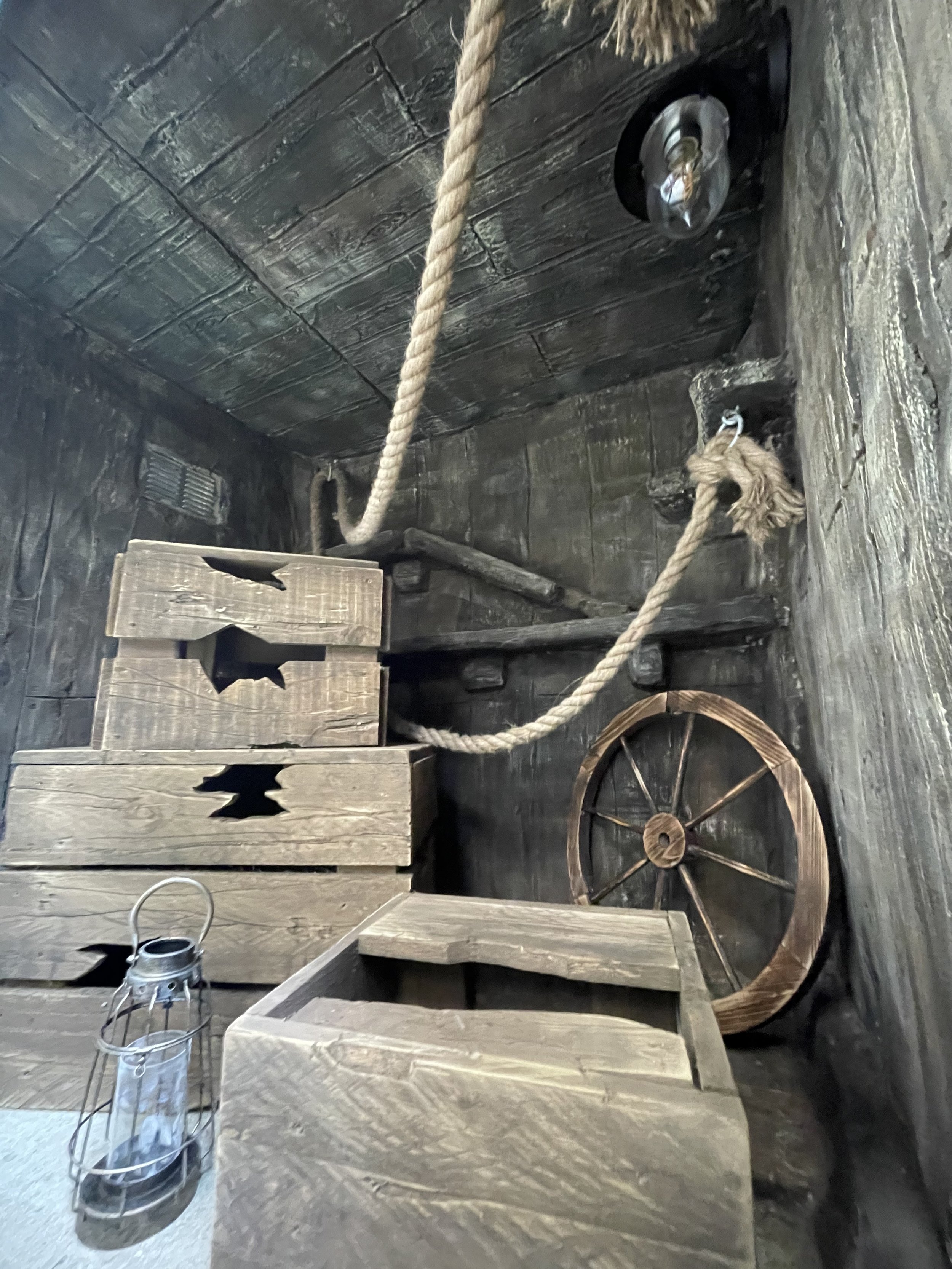 Rustic wooden crates, a metal lantern, a wooden wheel leaning against a dark wooden wall, a thick rope hanging from the ceiling, and wooden beams in a rustic setting.