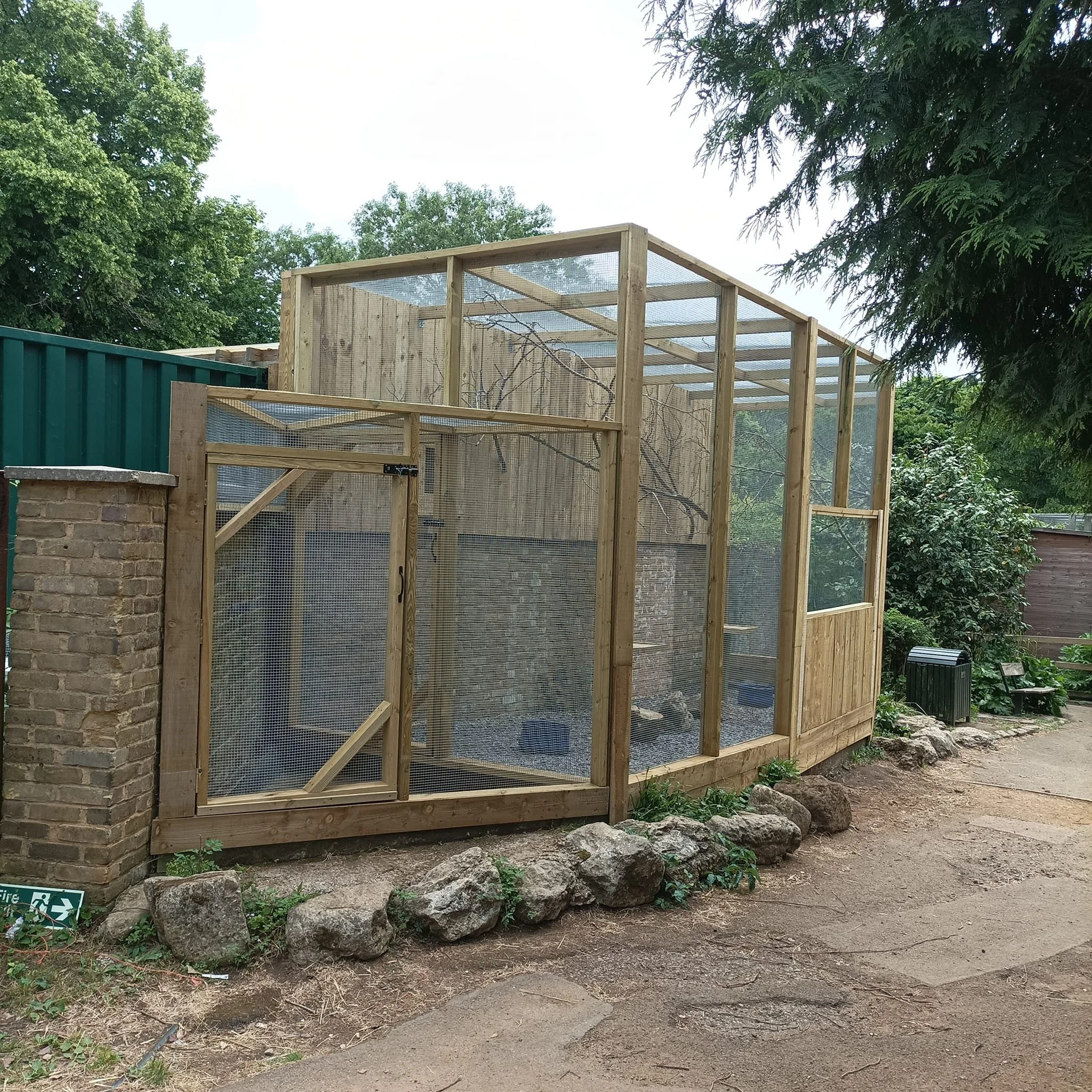 A wooden outdoor animal enclosure with wire mesh panels, situated next to a brick wall and surrounded by rocks and natural greenery.