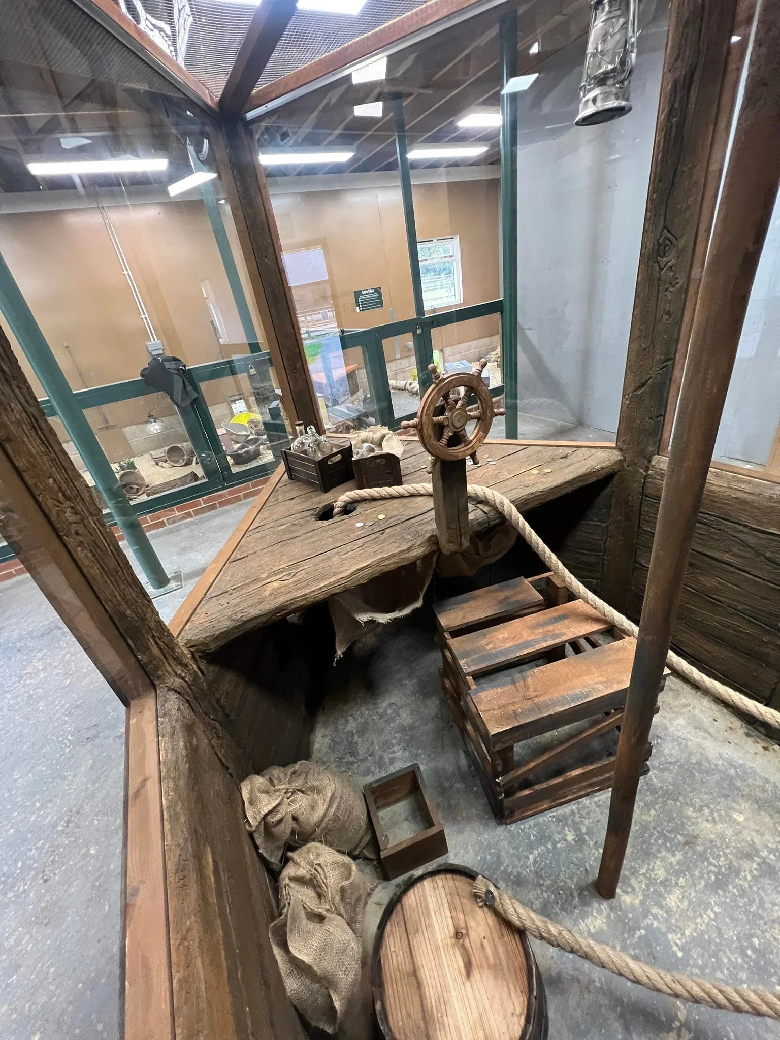 A nautical-themed museum exhibit with a wooden ship's wheel, a rope, burlap sacks, a wooden crate, and a barrel on a concrete floor, enclosed in a glass display case.