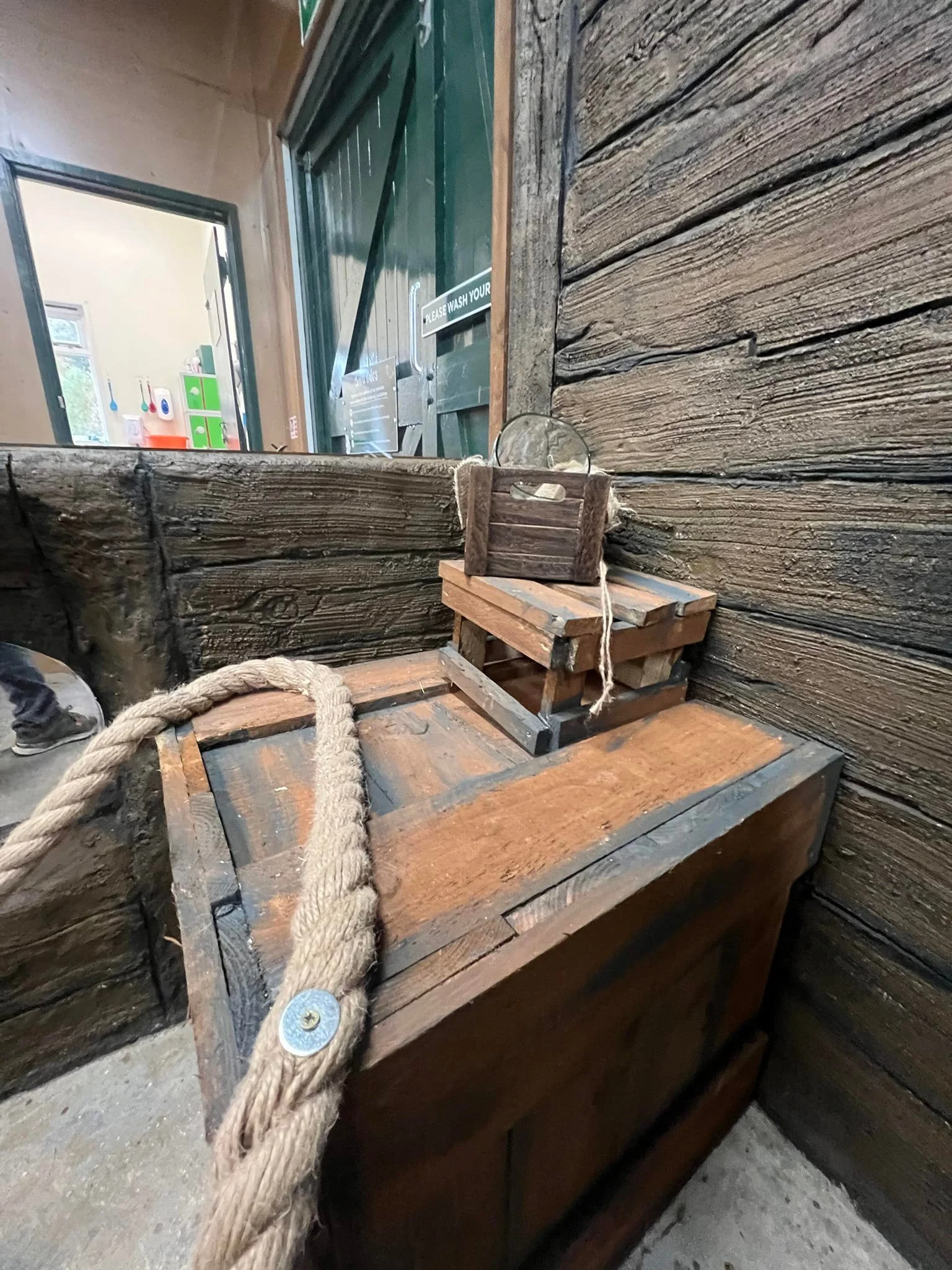 Small decorative display with a miniature wooden rowboat, a glass lens, and a thick rope on a wooden crate inside a rustic room with wooden walls.