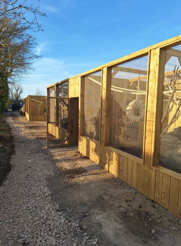 A wooden outdoor animal enclosure with wire mesh panels, situated along a dirt path under a blue sky, with trees in the background.