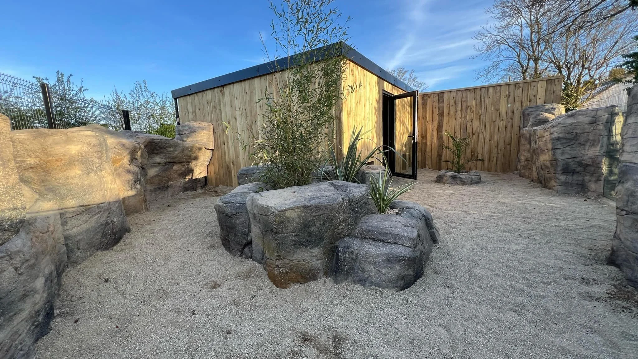 A small outdoor courtyard with large rocks, sand ground, and a wooden storage shed with a black roof. There are some plants and trees in the background under a blue sky.