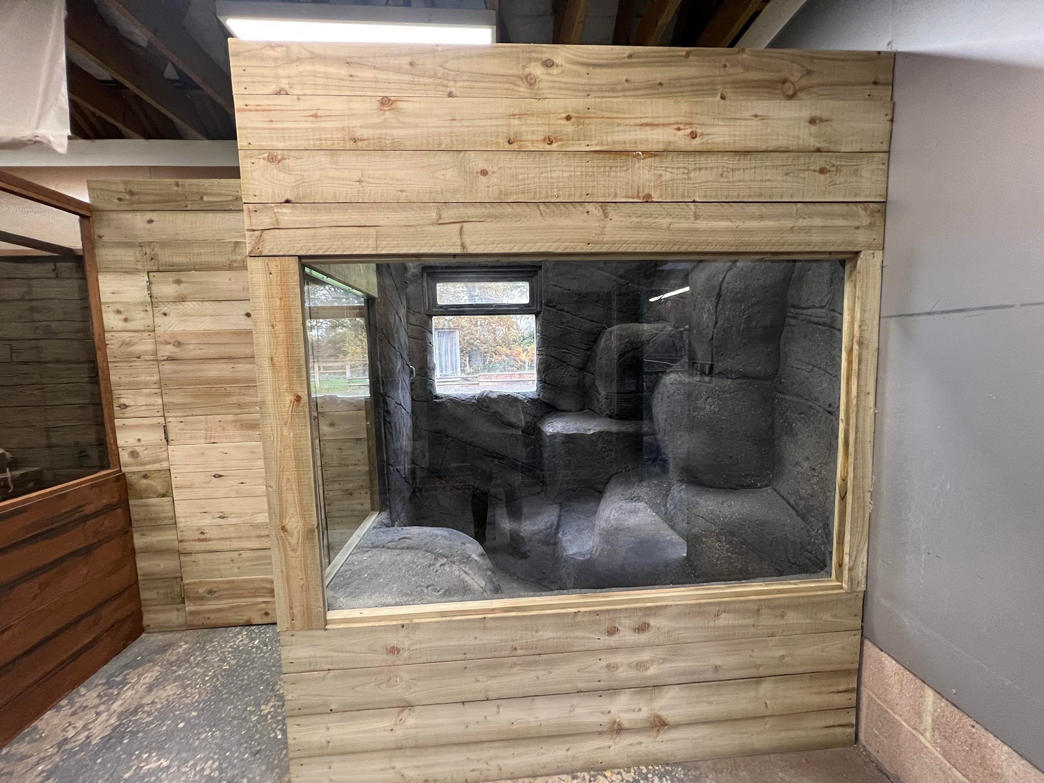 A display enclosure with a wooden frame and glass panel showing a rocky interior with gray stones and a small window in the background.