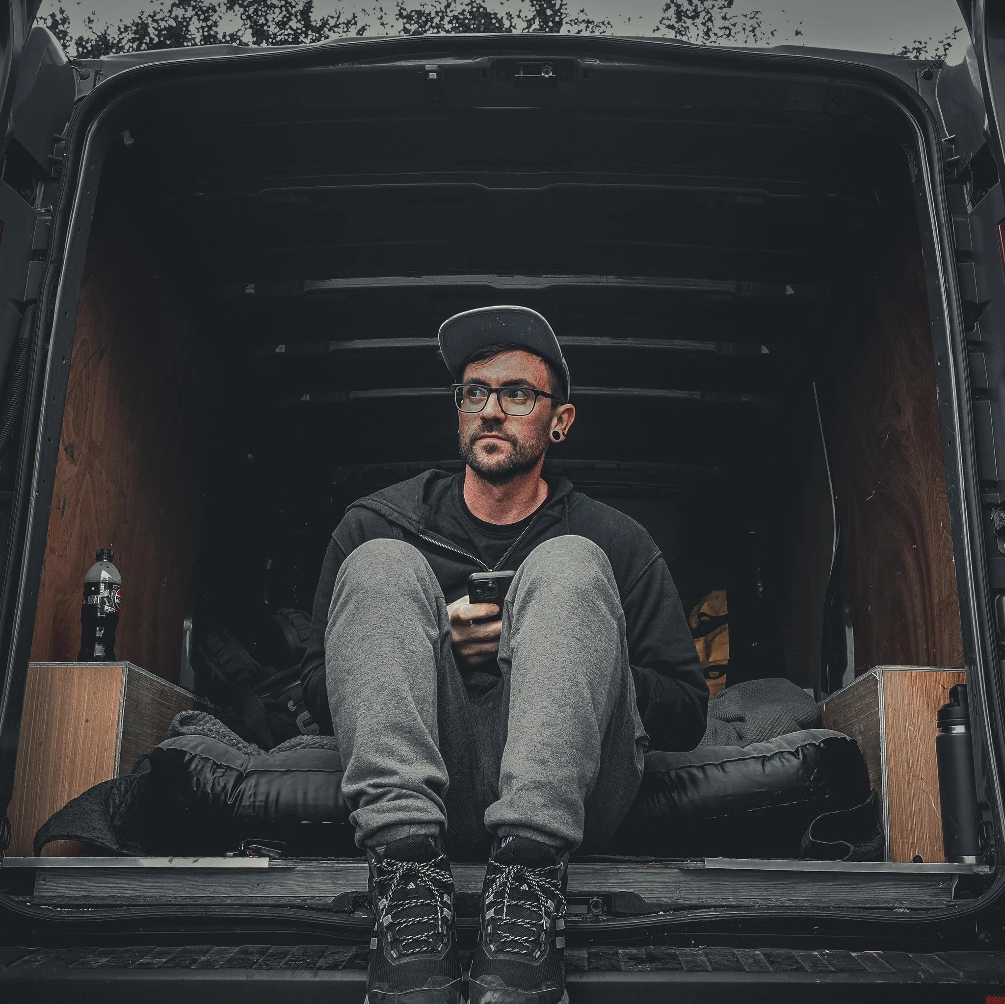 A man sitting inside the open back of a van. He is wearing glasses, a cap, a black hoodie, gray sweatpants, and hiking boots. He is looking to the side while holding a smartphone. Items like a water bottle, a backpack, and a sleeping bag are visible inside the van.