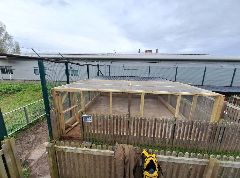 A fenced outdoor structure under construction with wooden framing, surrounded by a safety fence, with a small door and a sign, on a cloudy day.