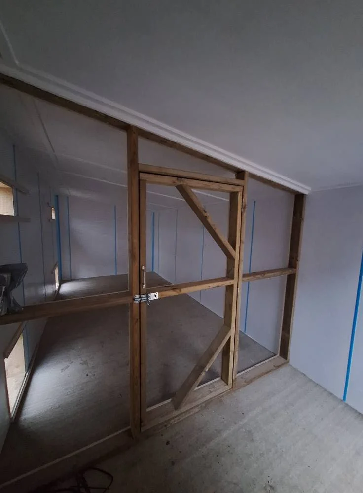 Interior of a room under construction with wooden framing for a wall or a door opening, with drywall partially installed, and a small window on the left side.