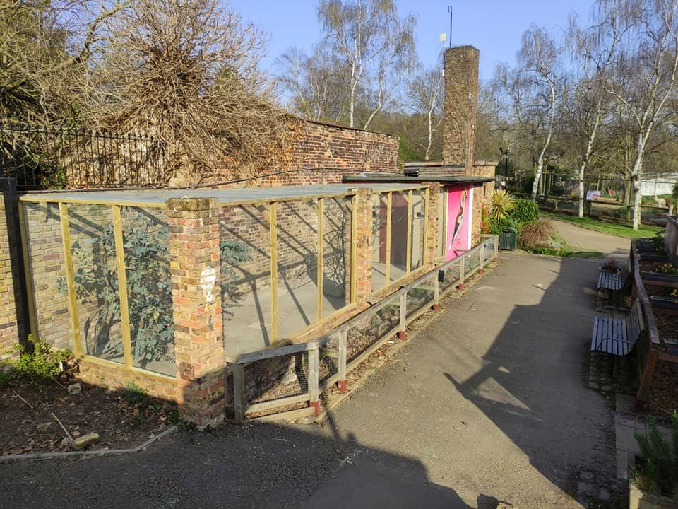 A small outdoor structure under construction with brick pillars, wooden framing, and plastic sheeting, next to a brick wall and a path in a park or garden area.