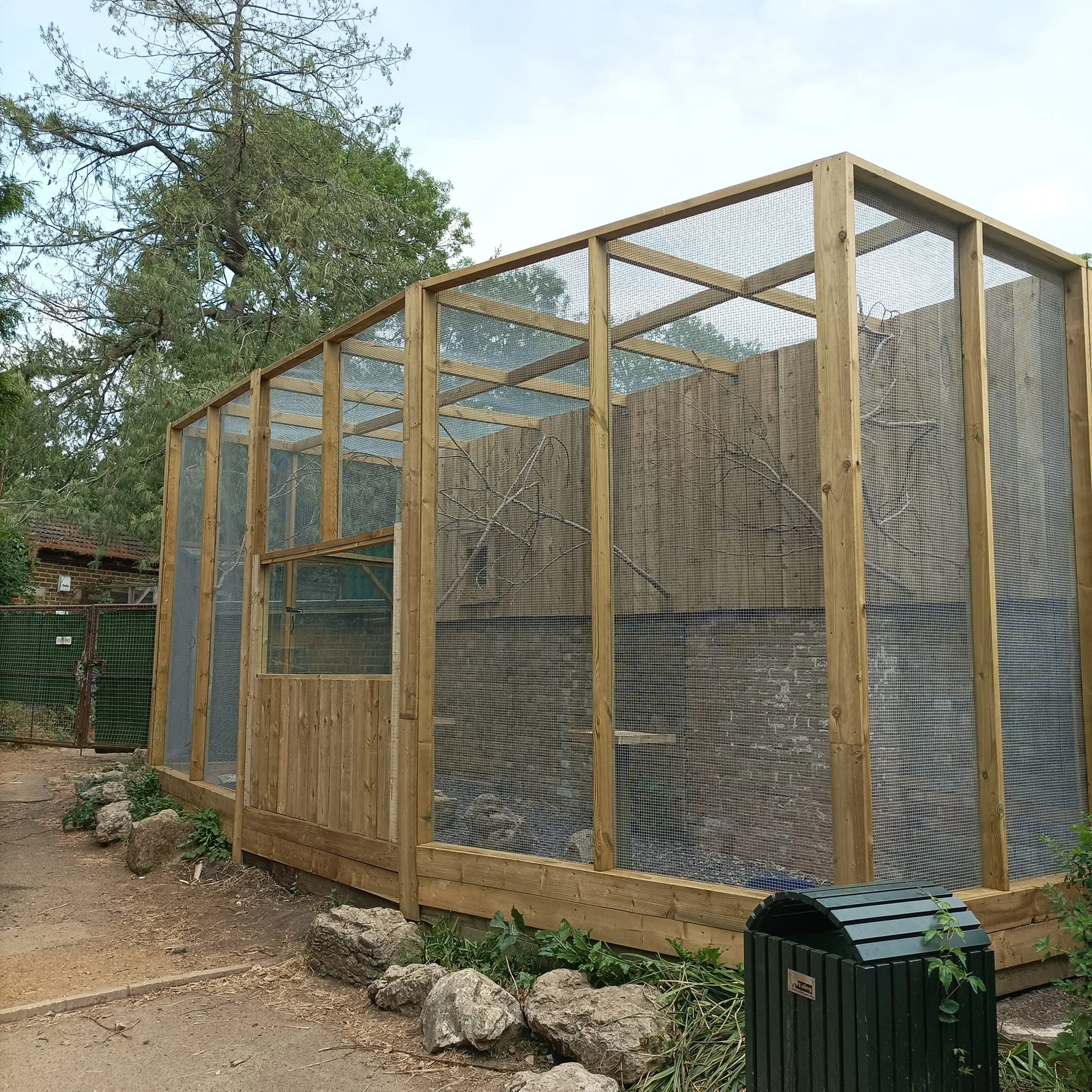 A wooden outdoor enclosure with mesh sides and roof, built on a stone foundation, situated in a park or zoo area.