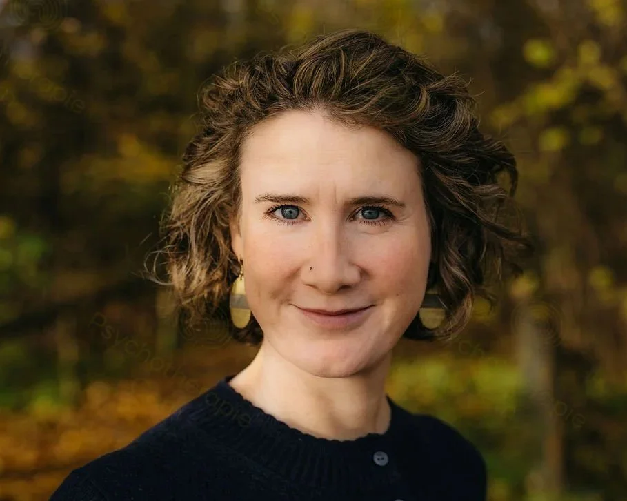 Porträt einer Frau mit kurzen, lockigen braunen Haaren vor einem herbstlichen Hintergrund, trägt schwarze Kleidung und goldene Ohrringe.