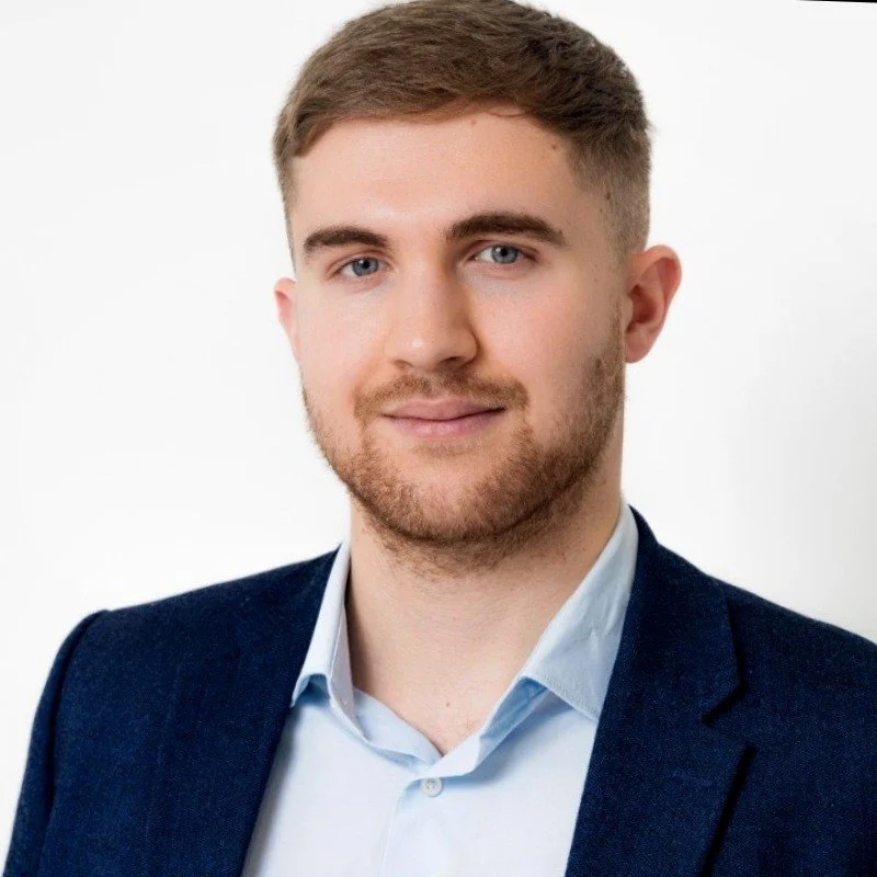 A young man with short light brown hair, blue eyes, and beard, dressed in a dark blazer and light blue shirt, posing against a plain white background.