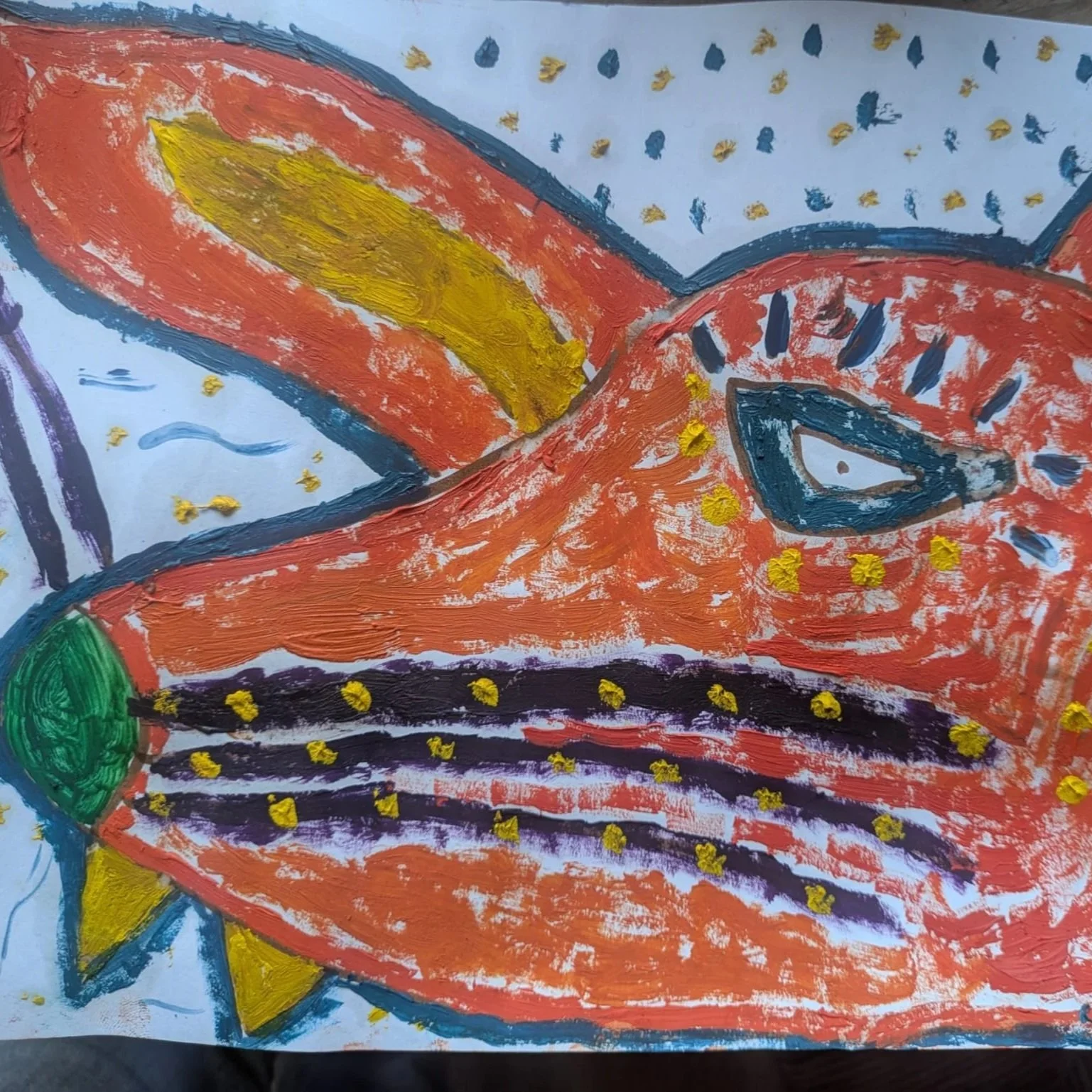 Children's drawing of an orange fish with black spot and a green tail, surrounded by blue and yellow feather-like shapes and dots, created with paint and markers.