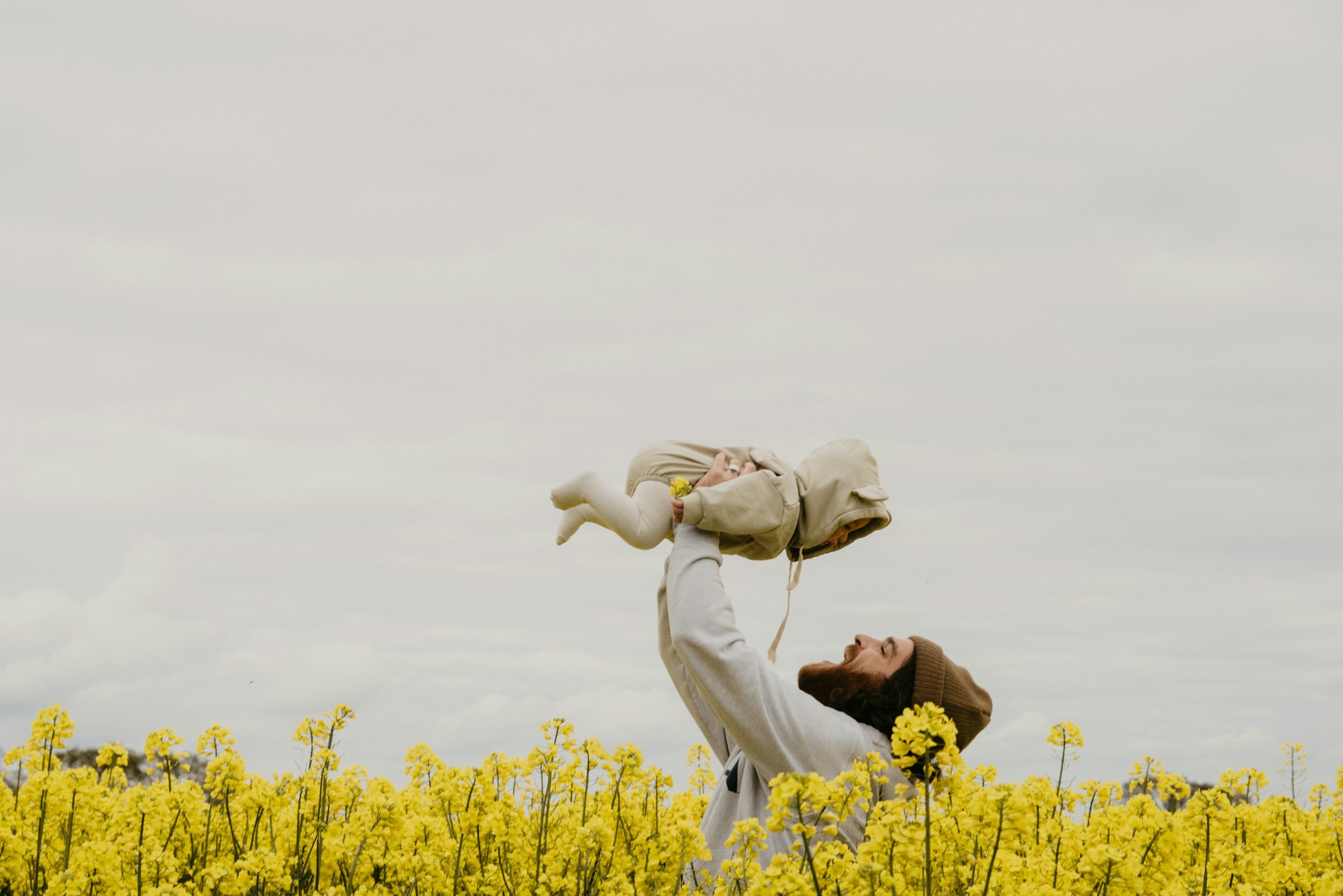 A man with a beard wearing a brown beanie and gray sweatshirt lifting a baby dressed in a beige jacket and white pants above his head in a yellow flower field on a cloudy day.