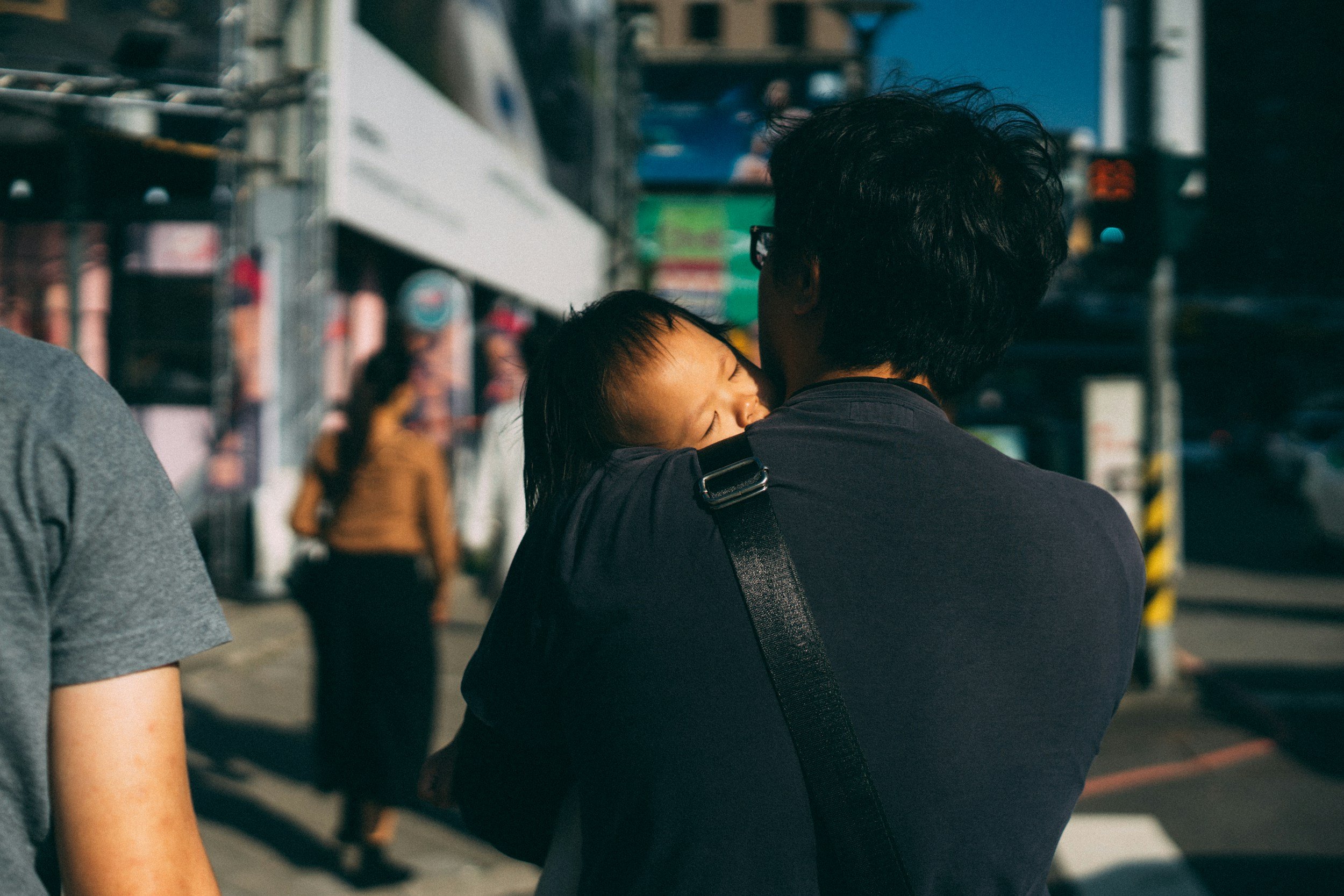 A person holding a sleeping child on their shoulder in an urban street scene with people and storefronts in the background.