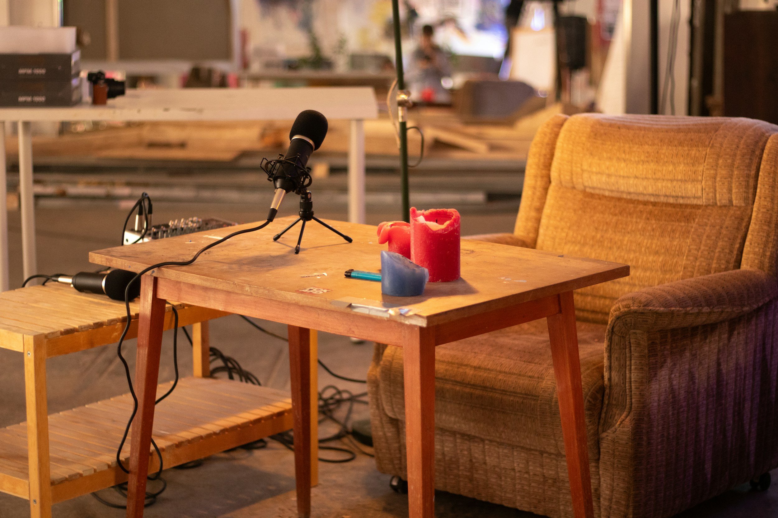 A cozy recording setup with a wooden table featuring two red candles, a blue candle, and a blue pen. There is a black microphone on a stand, a small mixing console, and exposed cables. An orange upholstered armchair is beside the table. In the background, there is a cluttered workspace with books, papers, and other objects.