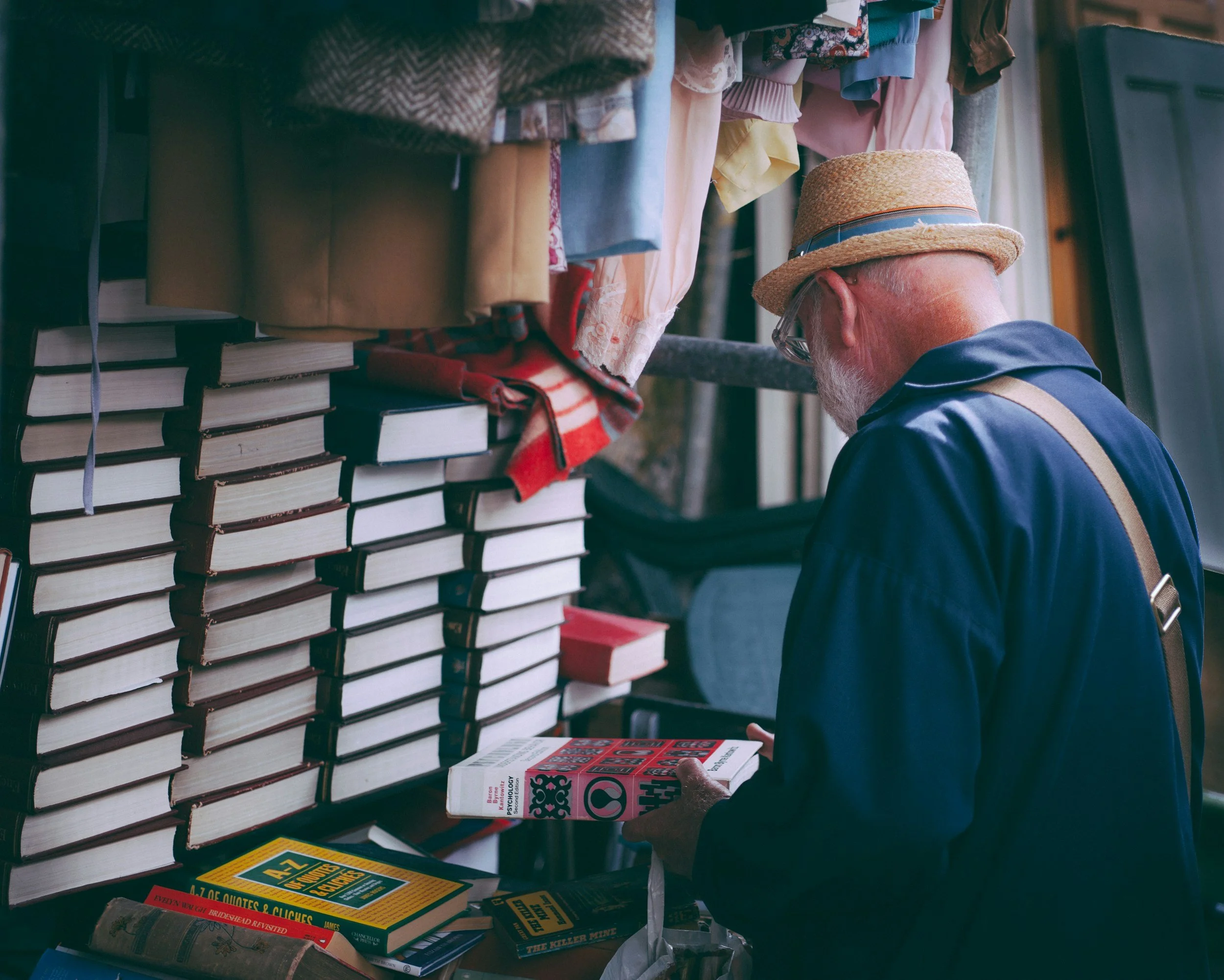 An elderly man with a white beard, wearing a straw hat and glasses, shopping at an outdoor book stall. He is looking at a book with a white cover that has a red and black design. The stall is filled with stacks of books, some on the table and others stacked in the background. There are also some clothing items hanging above the books.