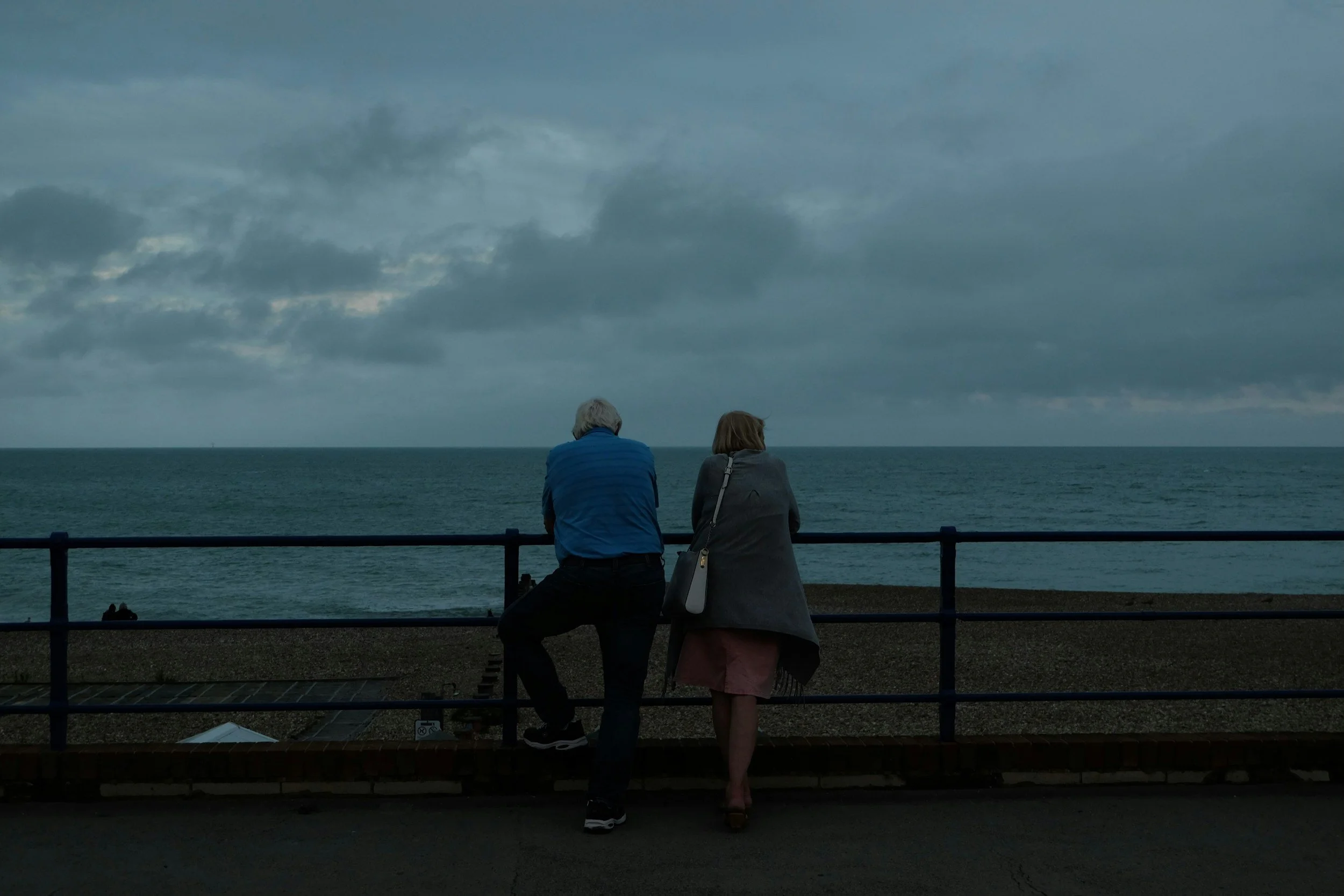 Two elderly people, a man and a woman, sit on a railing by the ocean on a cloudy day, looking out at the water.