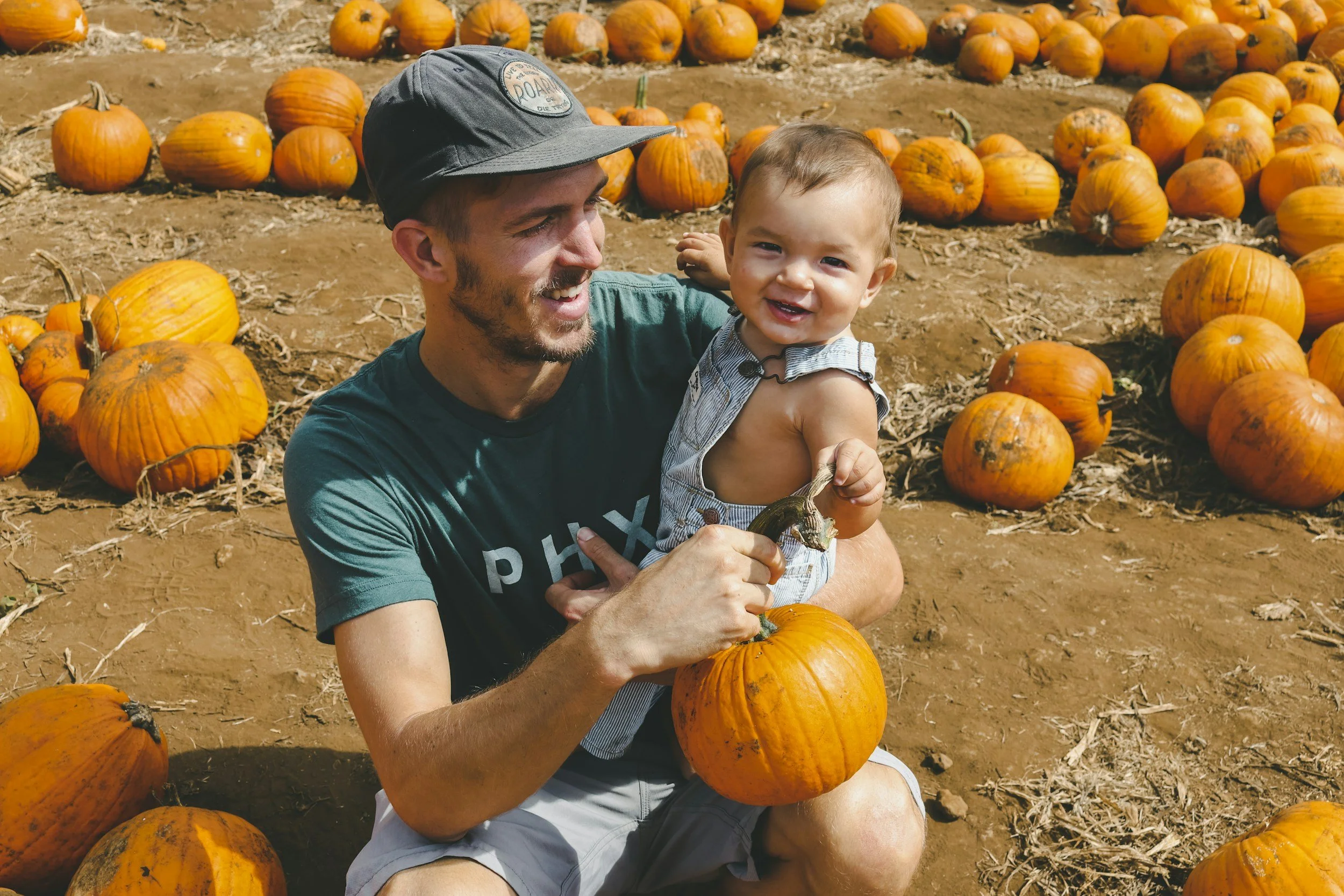 A man holding a smiling toddler in a pumpkin patch with numerous orange pumpkins on the ground and in the background.