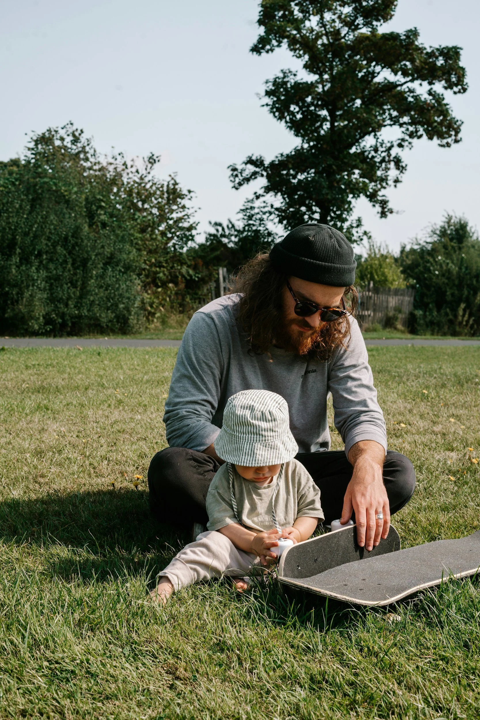 A man and a young child sitting on grass in a park, preparing to ride a skateboard. The man wears a gray long-sleeve shirt, black beanie, and sunglasses. The child wears a striped bucket hat and light-colored clothes, holding a small object.