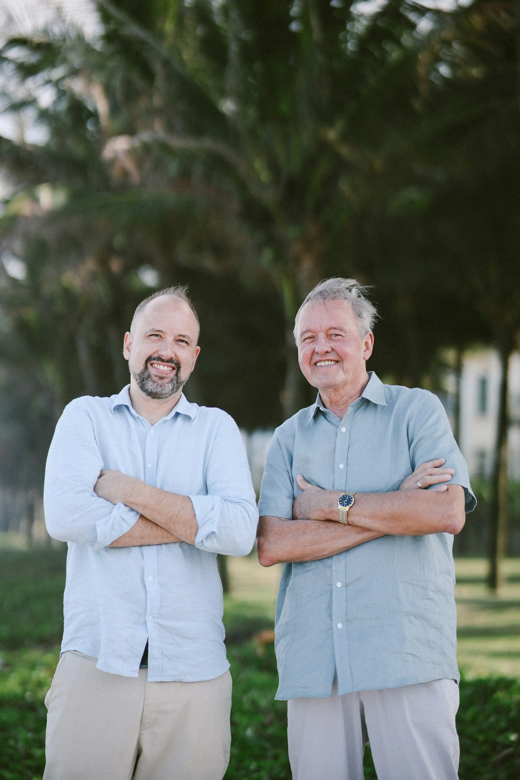 Two men smiling and crossing their arms outdoors in front of green trees.