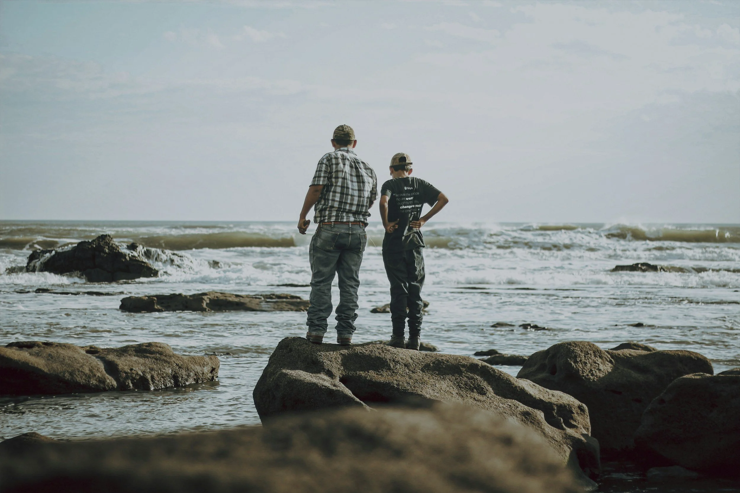 Two people standing on rocks at the beach, looking out over the ocean with waves and cloudy sky in the background.
