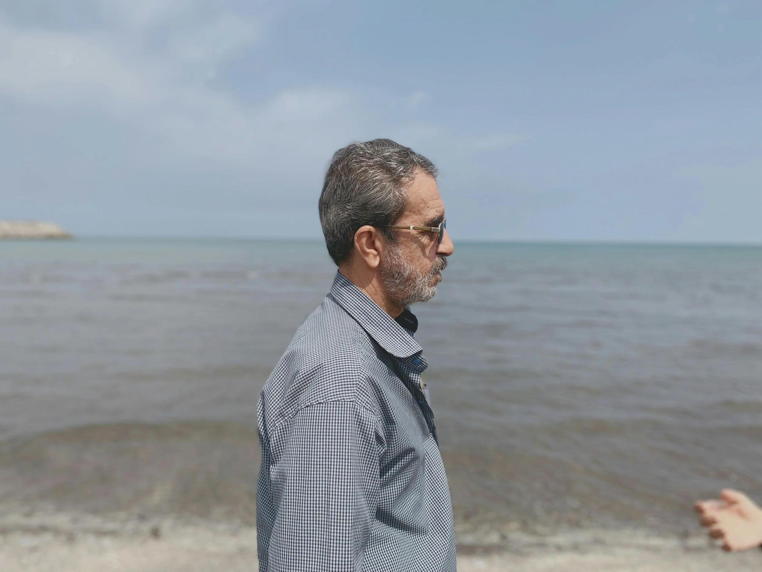 Side view of a middle-aged man with gray hair and beard, wearing glasses and a checkered shirt, standing on a beach near the sea with his eyes closed.