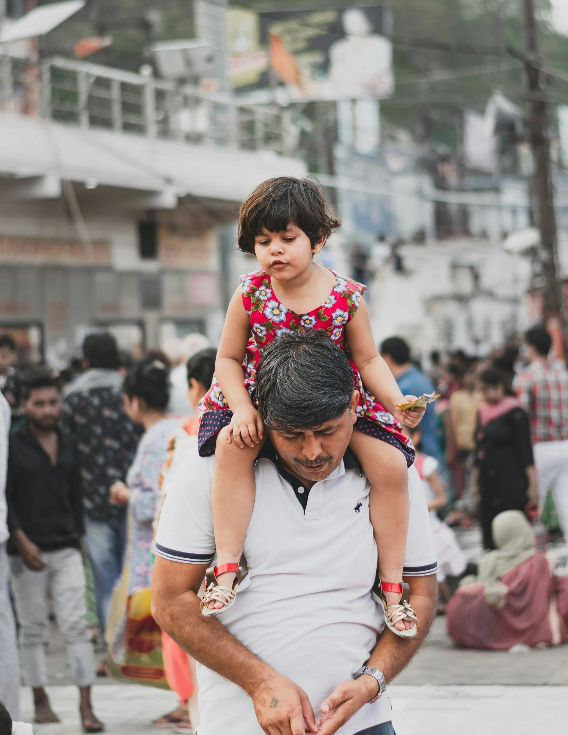 A man carrying a young girl on his shoulders in a busy outdoor market or street scene.