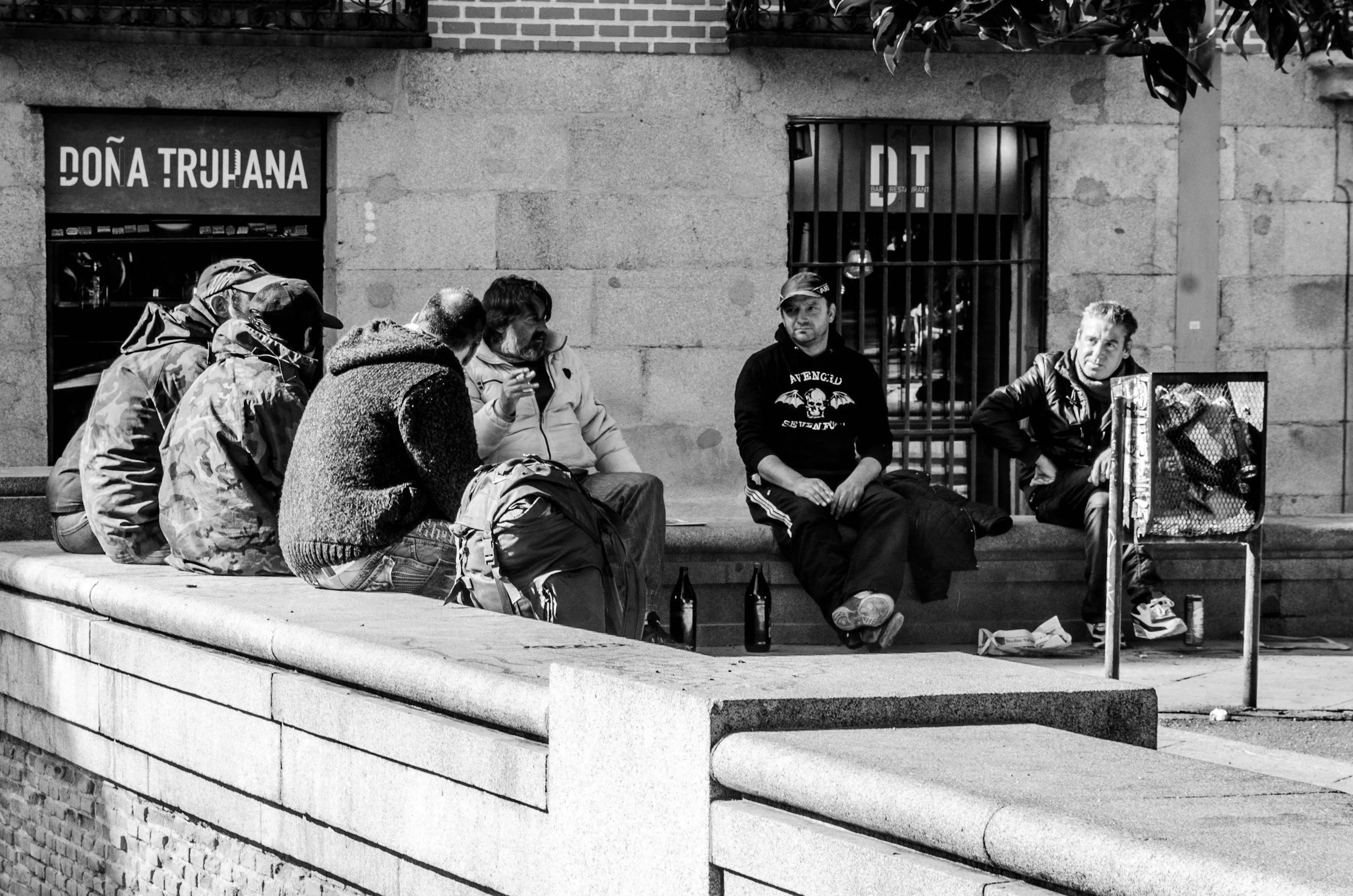 Seven men sitting on a stone ledge outside a building, some smoking and some with bottles, with a shopping cart nearby.