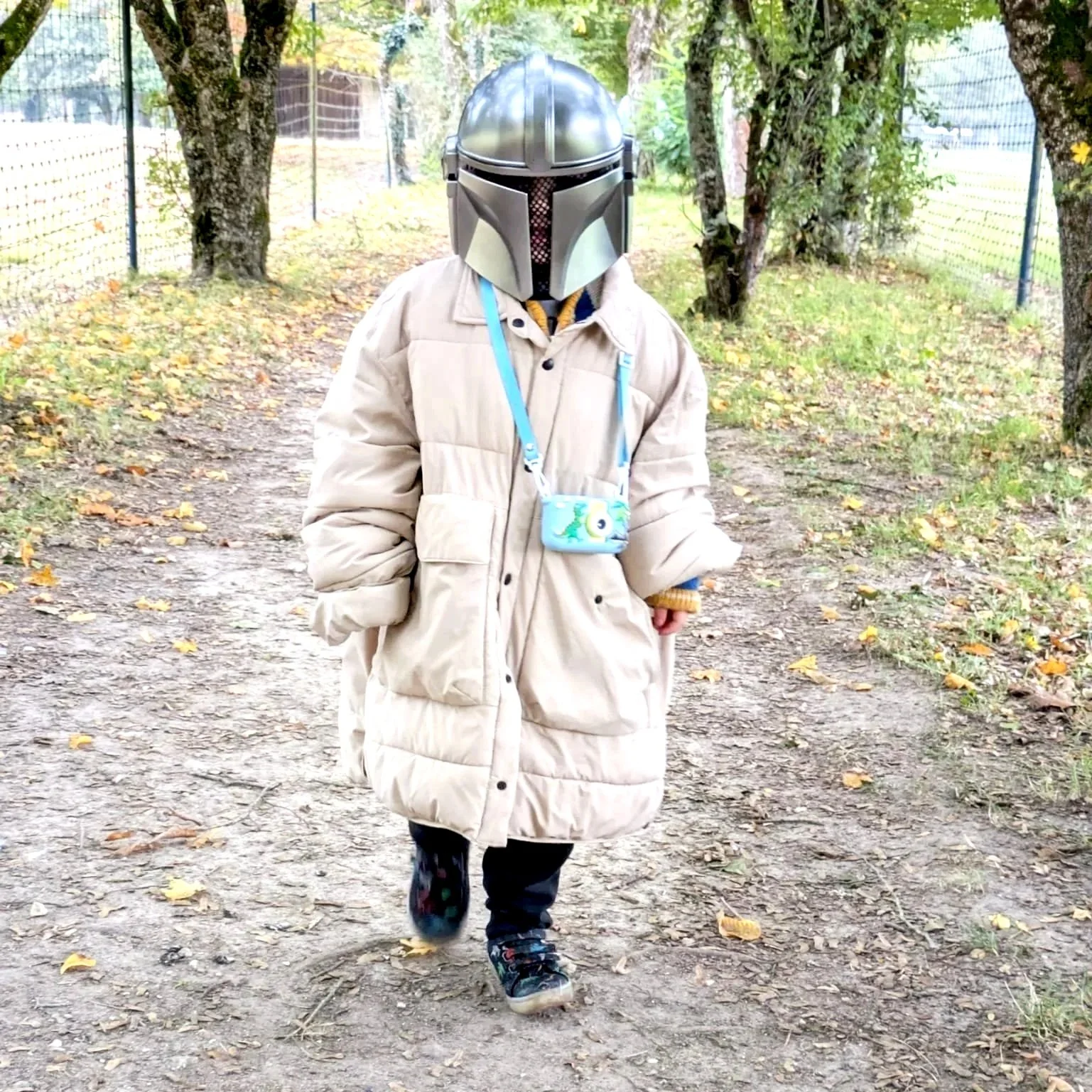 Child walking outdoors on a dirt path, wearing a beige puffer coat, colorful sneakers, and a silver helmet resembling Star Wars Mandalorian helmet, with a small blue camera or bag hanging around their neck, in a wooded area with trees and a fence.