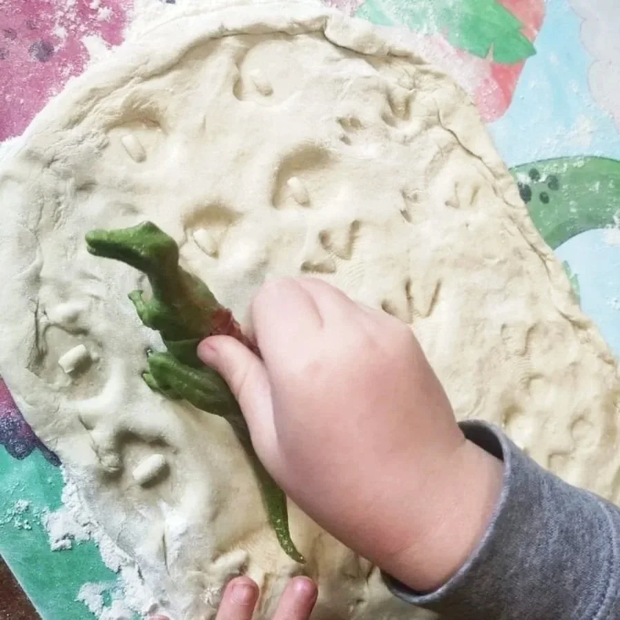 A child is pressing a green chili pepper into a flattened piece of dough with face impressions. The dough appears to be for baking.