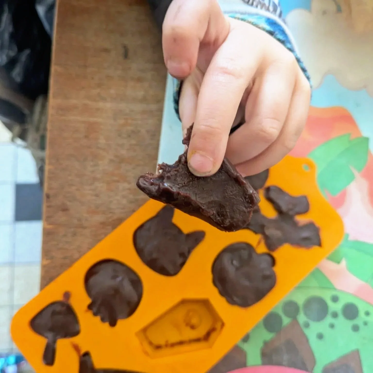 Close-up of a child's hand holding a chocolate treat, with a colorful silicone mold and a decorated table in the background.