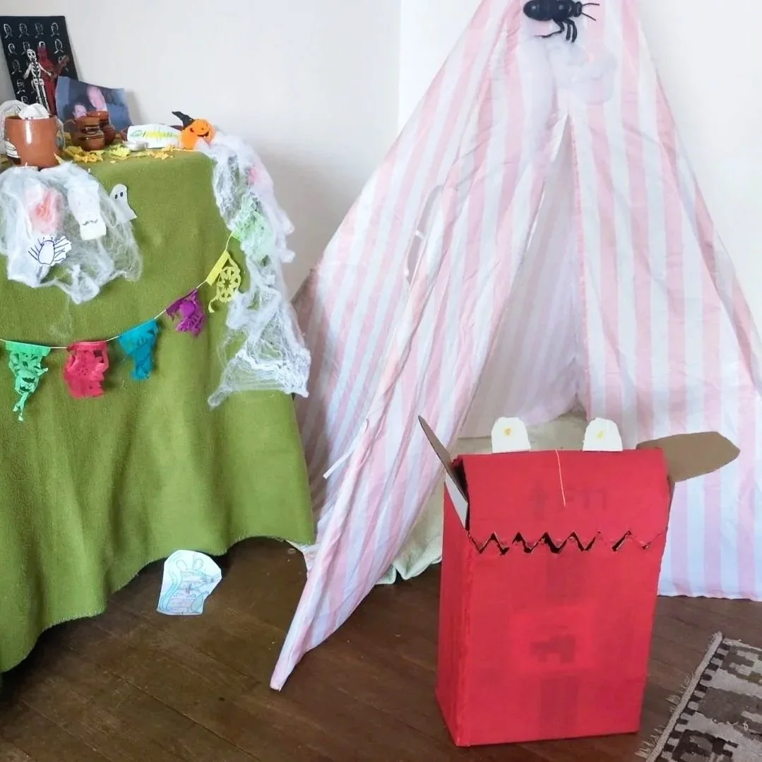 A Halloween-themed display with a green table covered in white tulle and Halloween decorations, next to a pink and white striped fabric tent, and a red cardboard box designed to look like a monster's mouth with paper eyes and cardboard teeth.