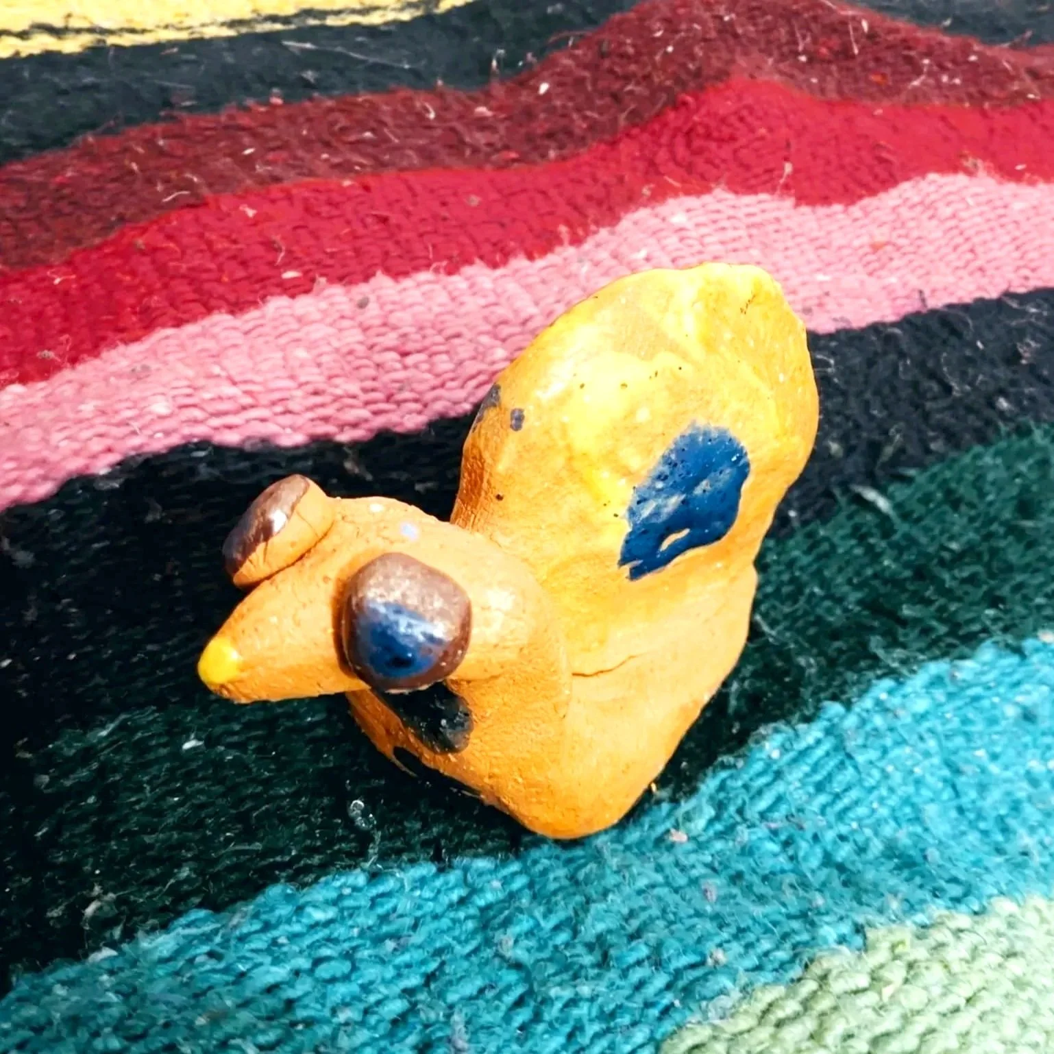 A small yellow rock with blue spots and black accents, shaped like a duck, resting on a multicolored woven fabric.