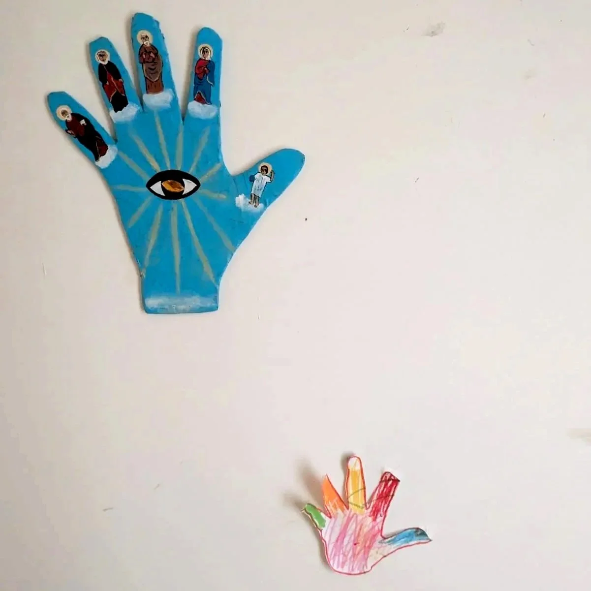 Colorful paper hand cutouts representing religious imagery, with a large blue hand featuring saint figures and an eye symbol, and a smaller rainbow-colored hand at the bottom.